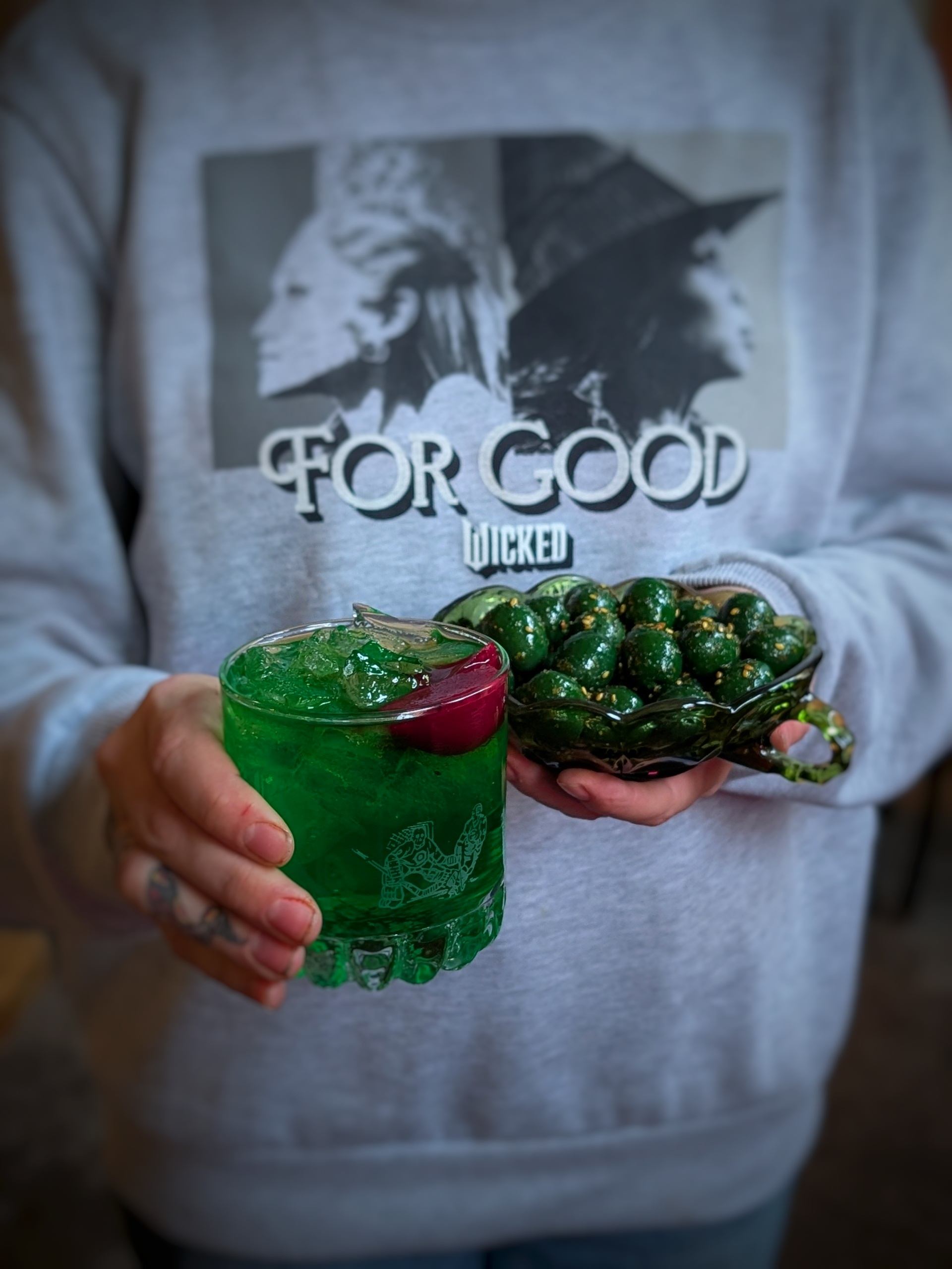 Person holding green drink and snack, wearing Wicked-themed sweatshirt with Elphaba and Glinda.
