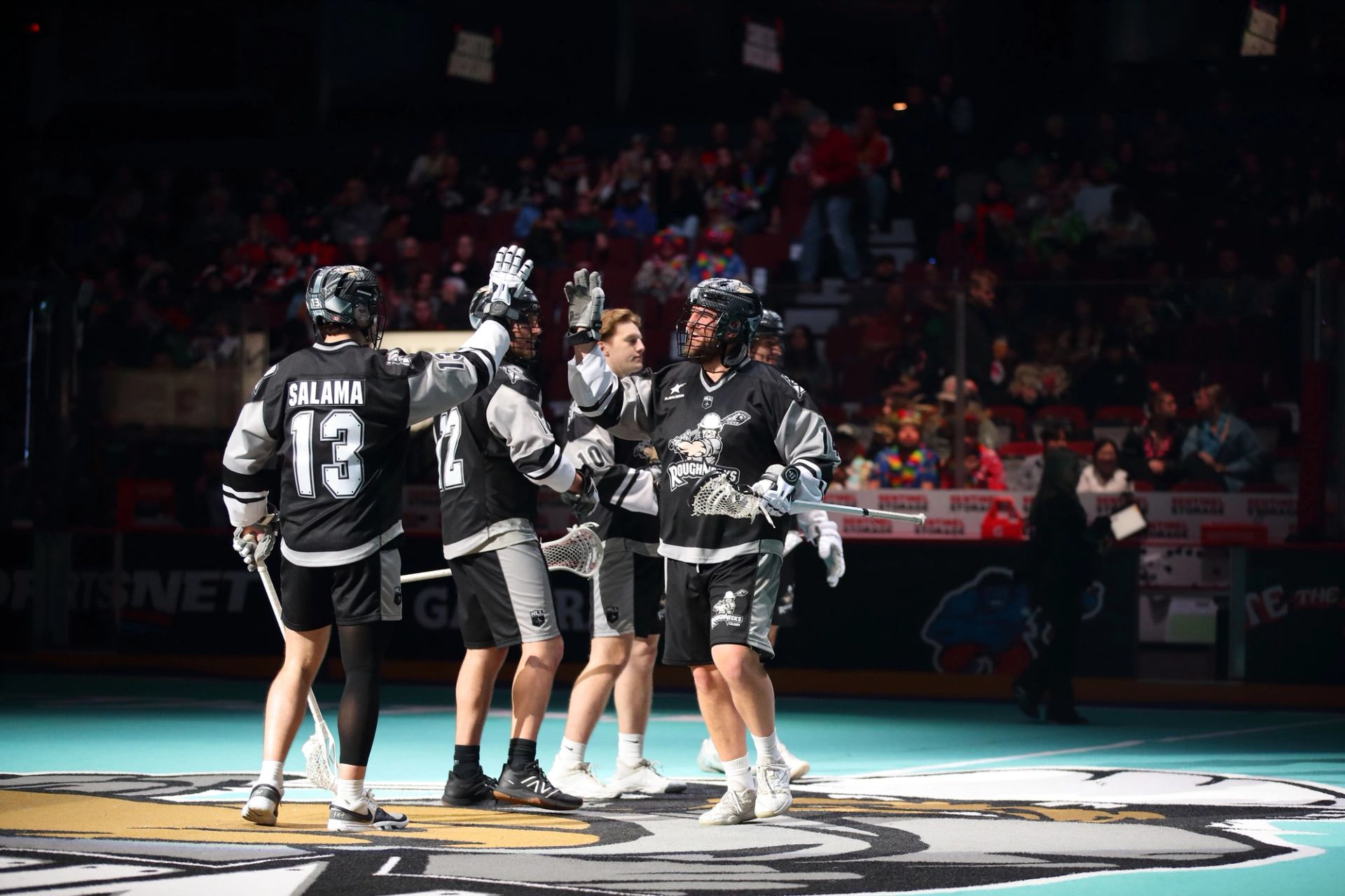 Lacrosse players in black and white uniforms, on indoor field with crowd behind.