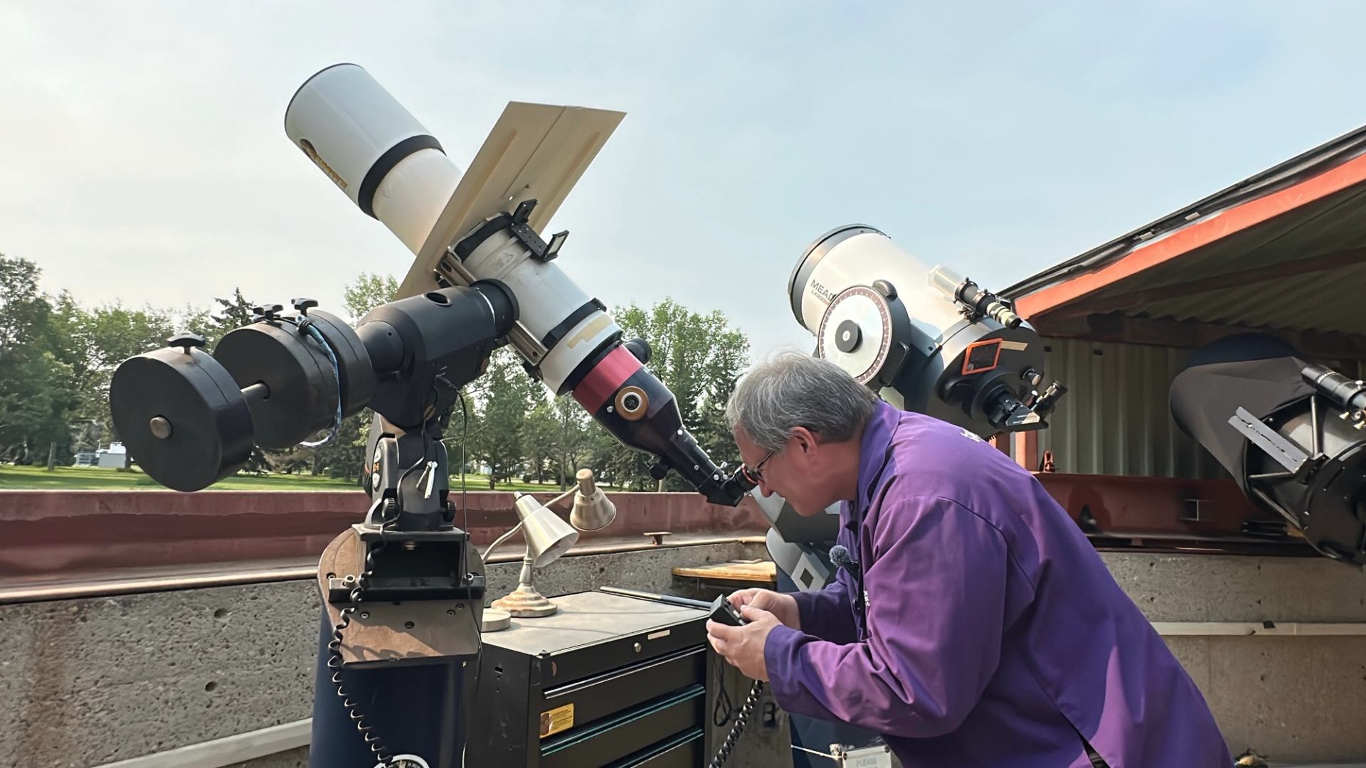 Astronomer in purple shirt looks through large outdoor telescope beside observatory equipment.