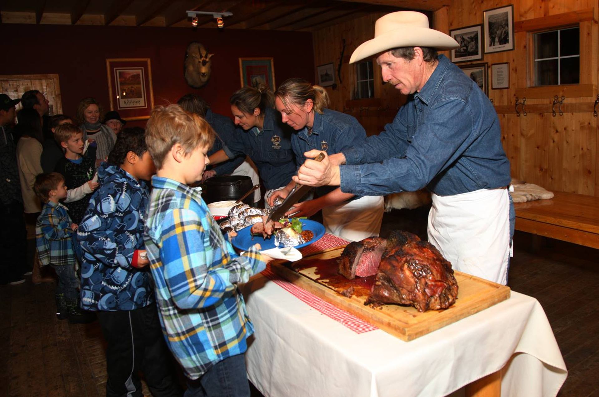 People serve food at rustic hall with cowboy hats and long table of roast meat.