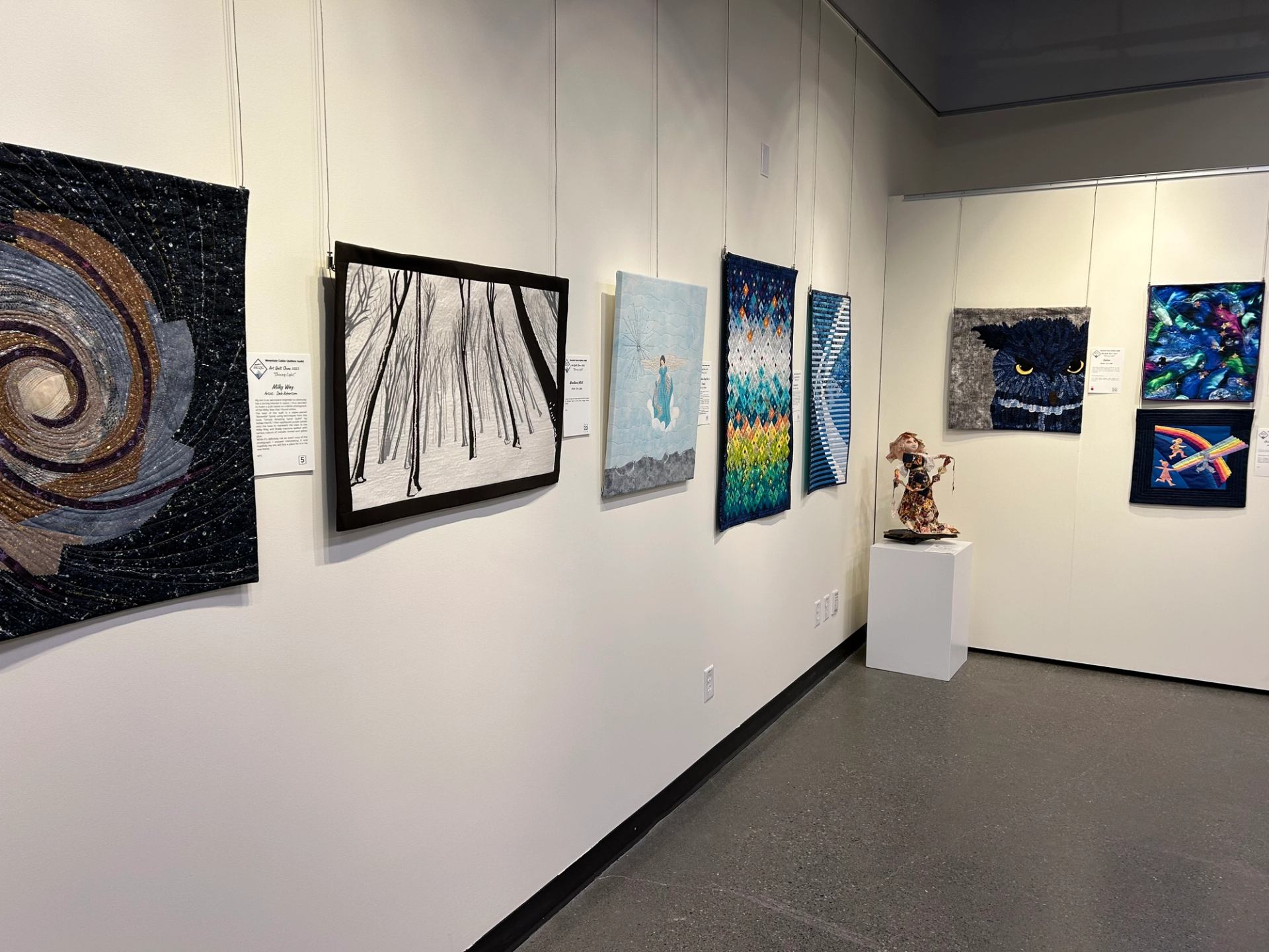Art quilt gallery display with multiple framed textiles and a small sculpted figure on a pedestal.