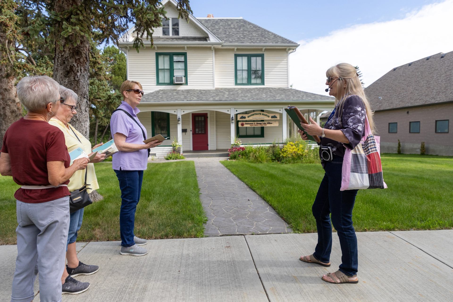 Tour guide with headset speaks to three people holding booklets outside historic house.