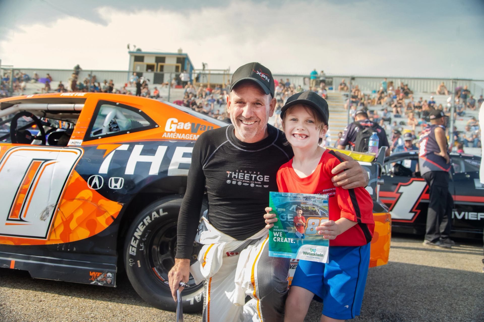 Driver kneeling next to a young fan holding a program in front of an orange NASCAR car on race day.