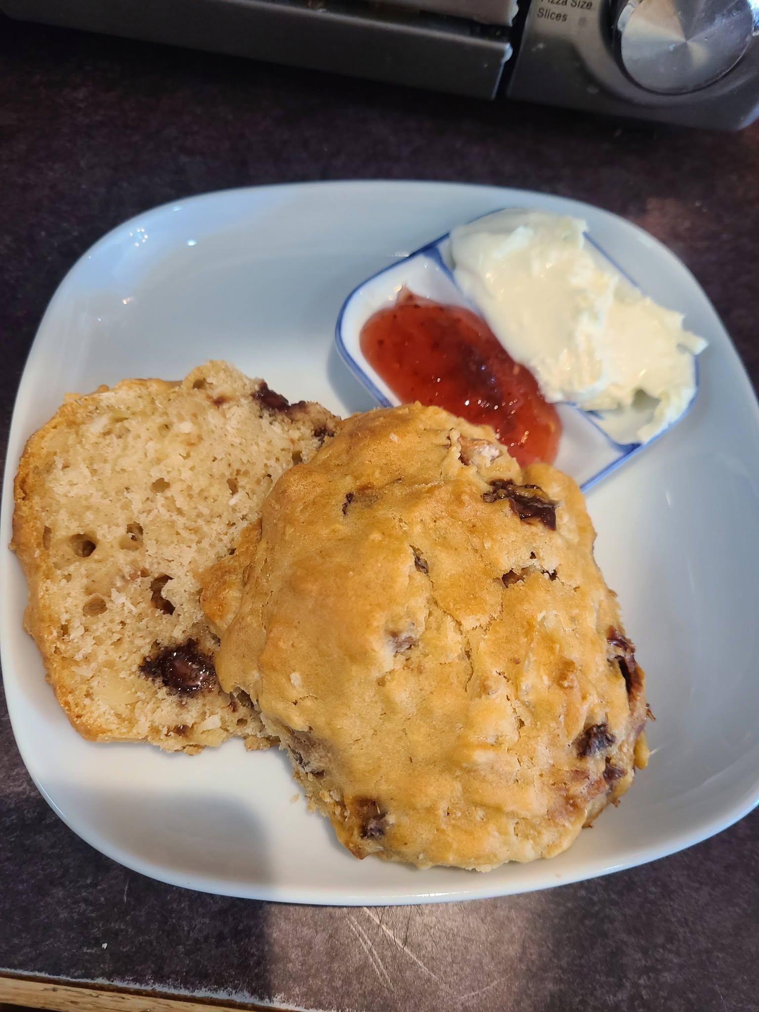 A plate holds a sliced raisin scone with strawberry jam and cream beside it for serving.