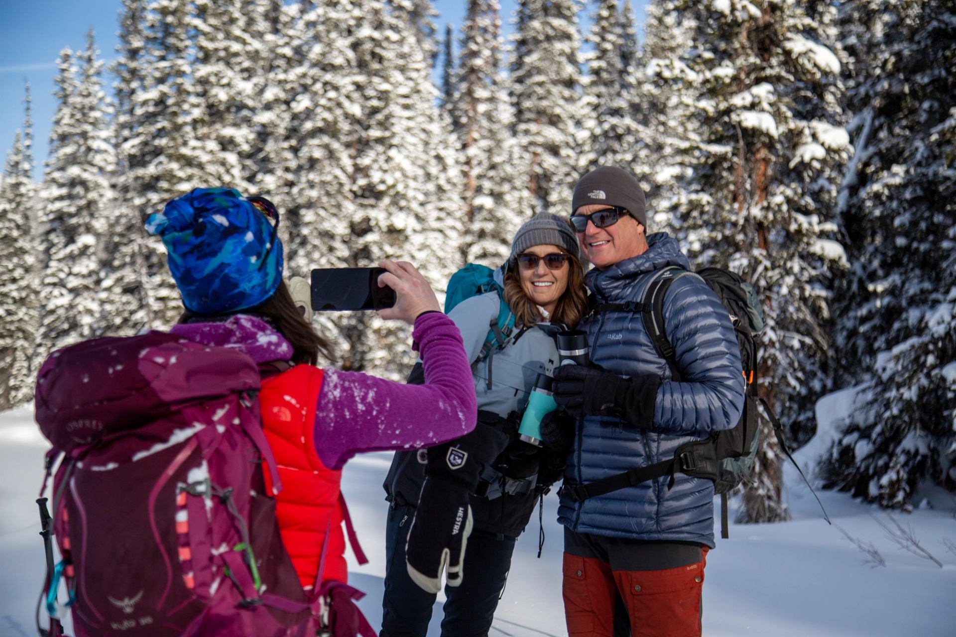 Person photographing two others posing on a snowy trail lined with pine trees