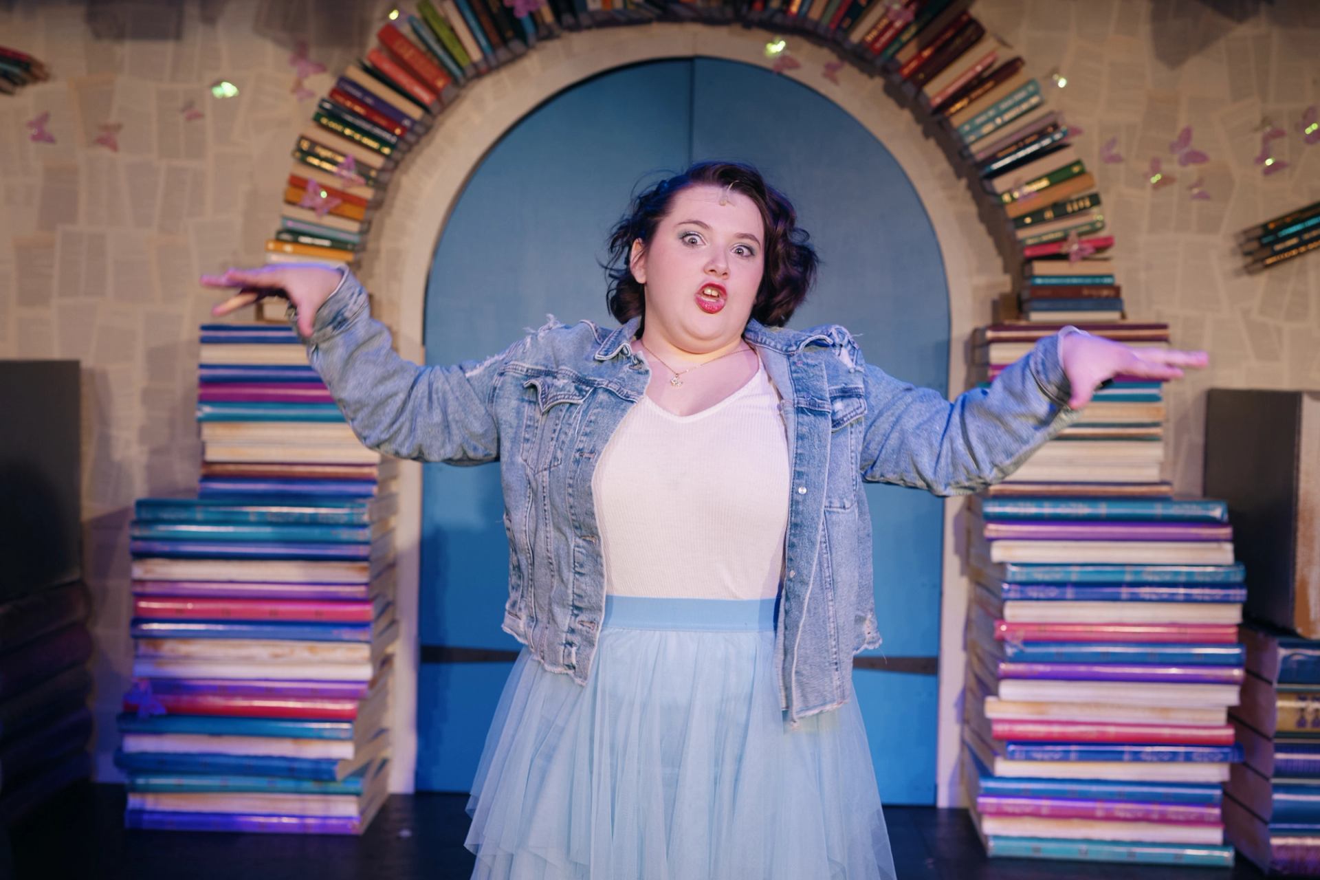 Performer in denim jacket and white dress on stage, arms out, surrounded by stacked books.