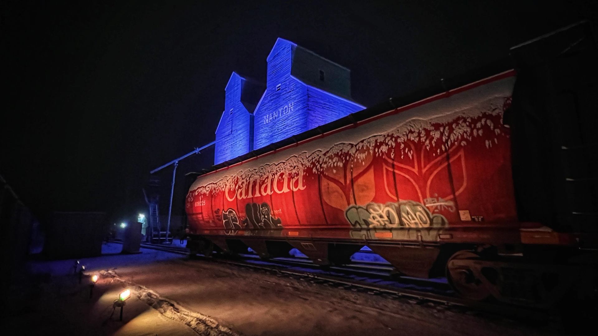 Blue-lit grain elevators behind a red rail car on a winter night.