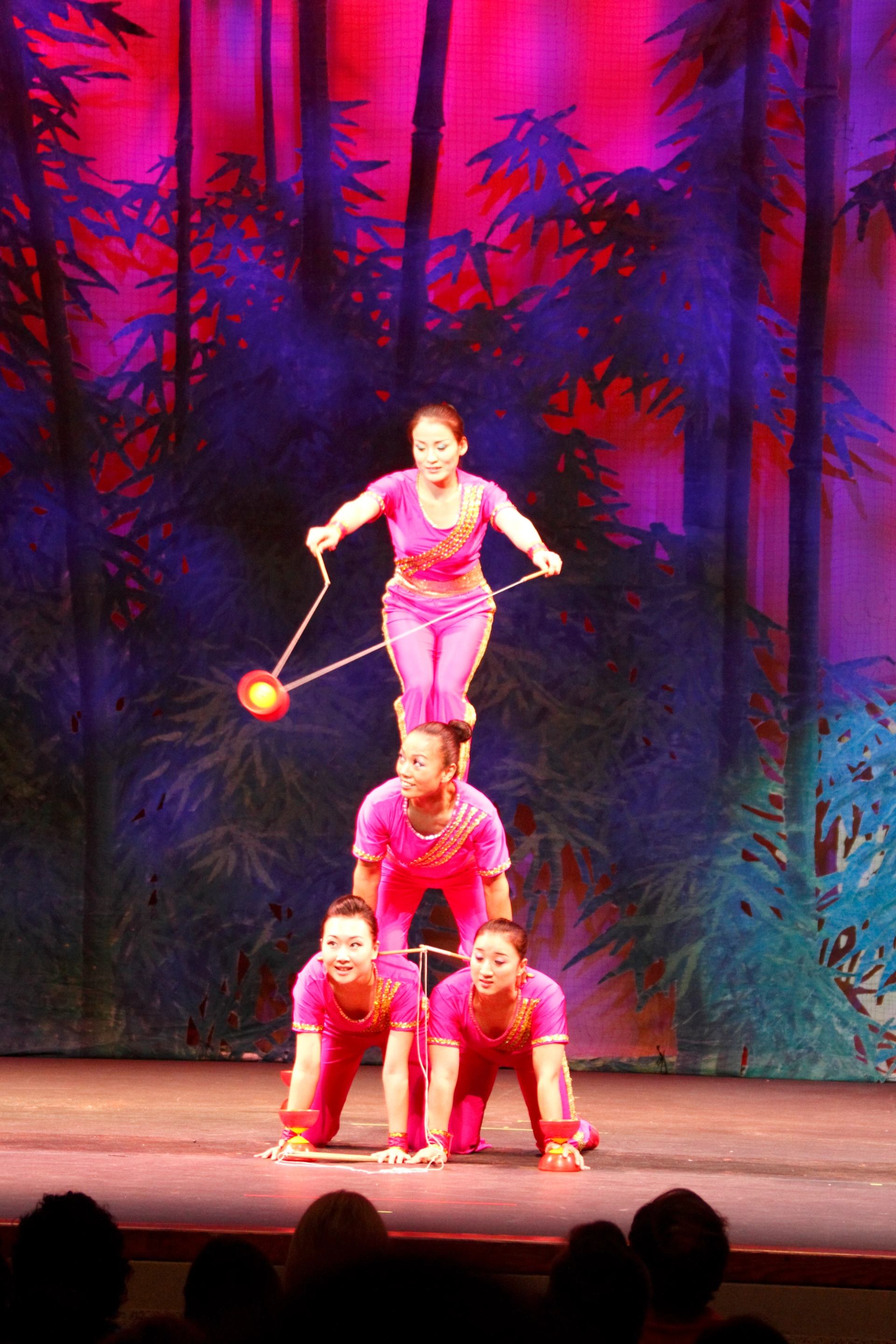 Acrobats in pink costumes balance in a human pyramid while spinning a diabolo.