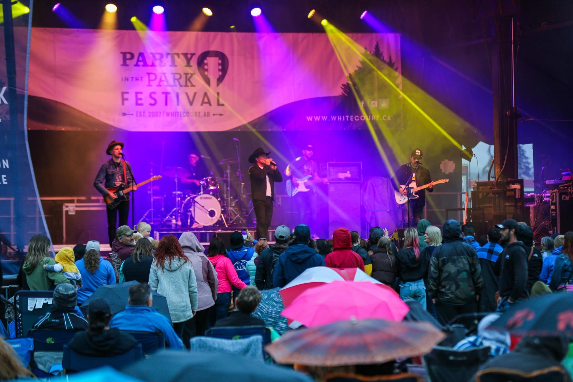 Band performing onstage at Party in the Park as a crowd watches in the rain.