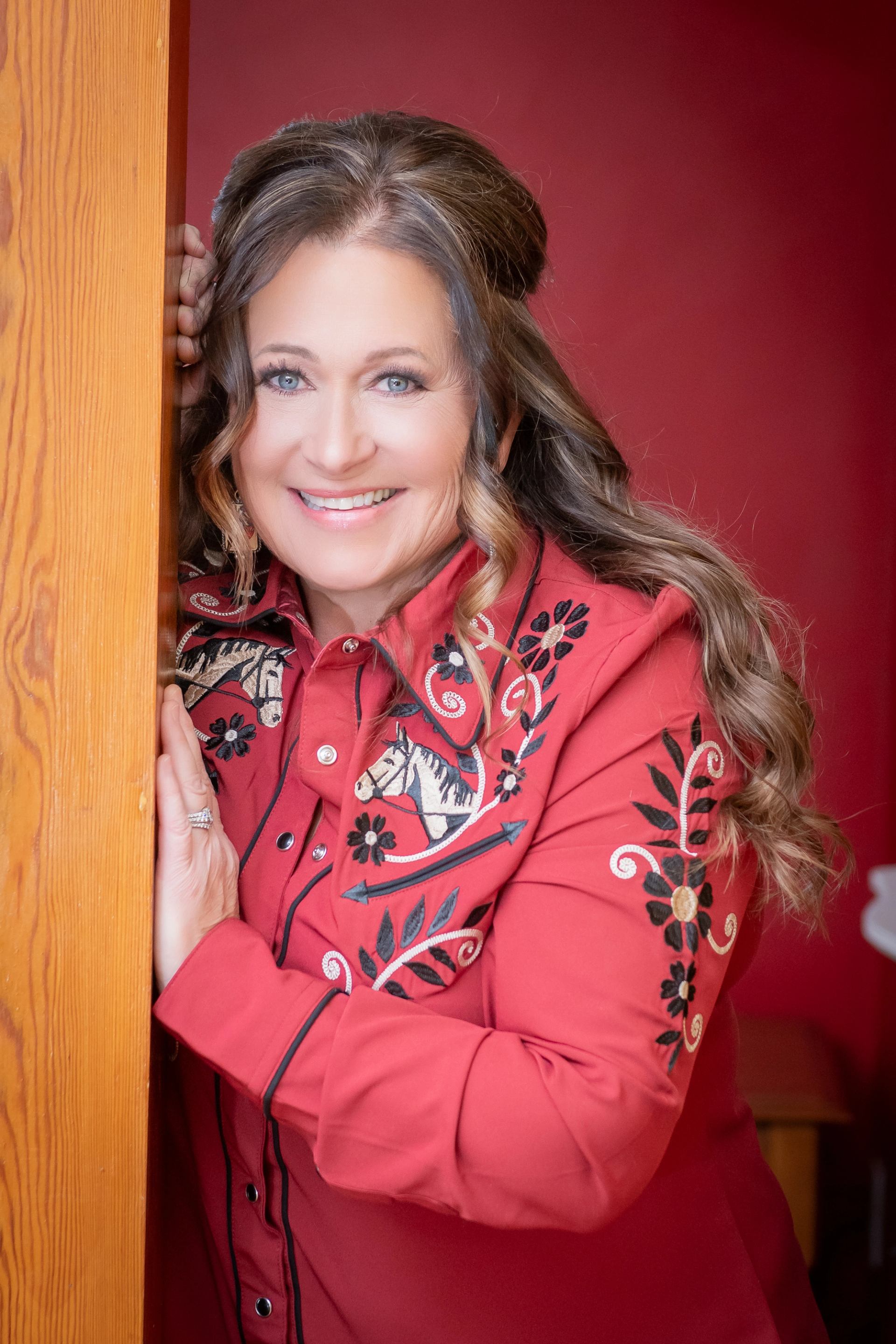 Woman in embroidered red western shirt leans on wooden post against red wall.