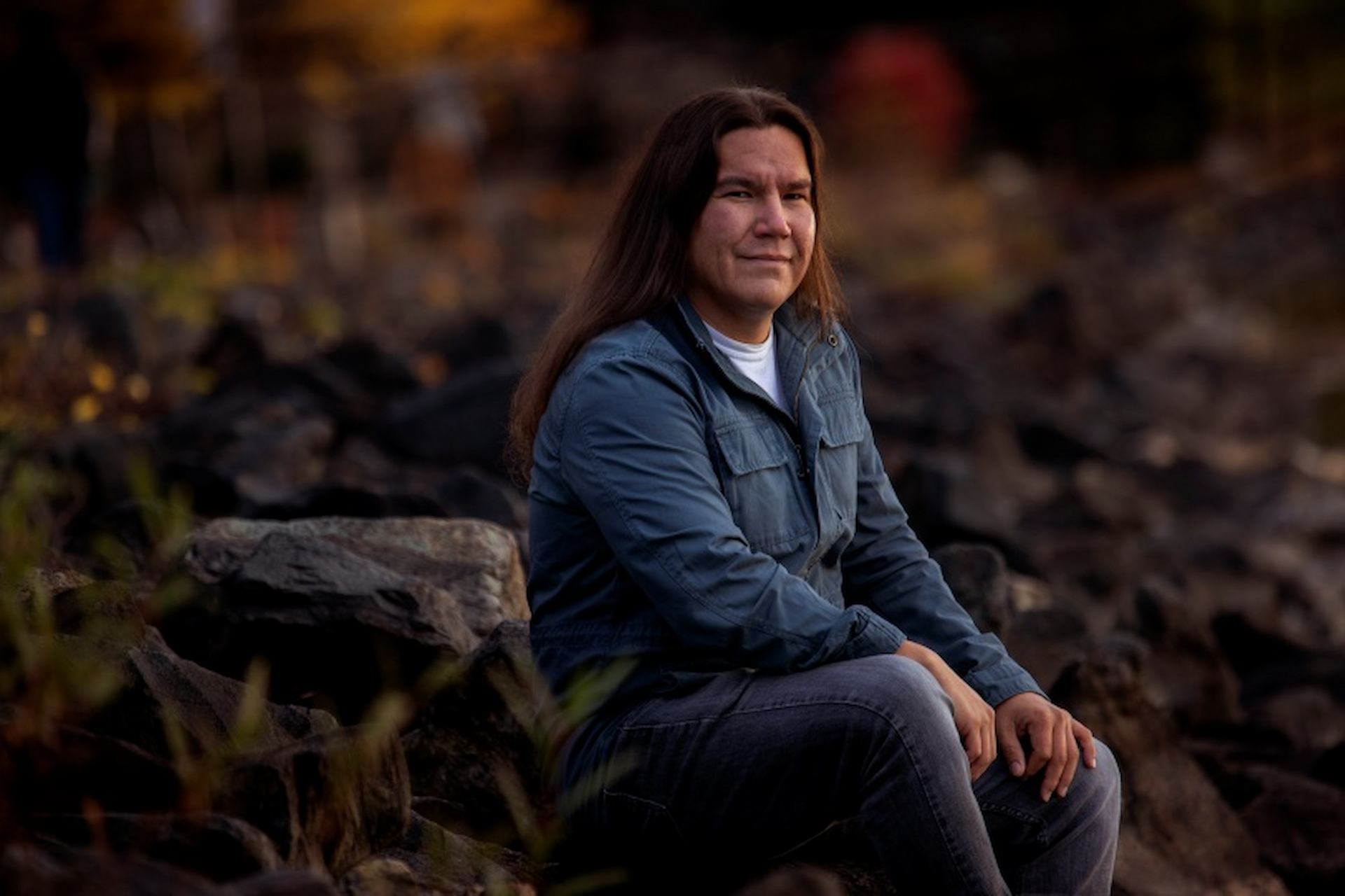 A person with long hair dressed in a blue jacket and jeans sits on large rocks outdoors, smiling slightly.