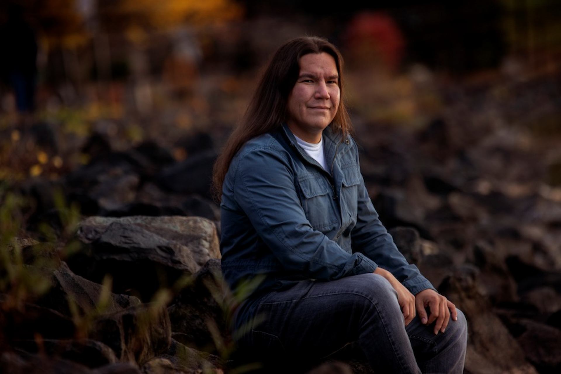 A person with long hair dressed in a blue jacket and jeans sits on large rocks outdoors, smiling slightly.