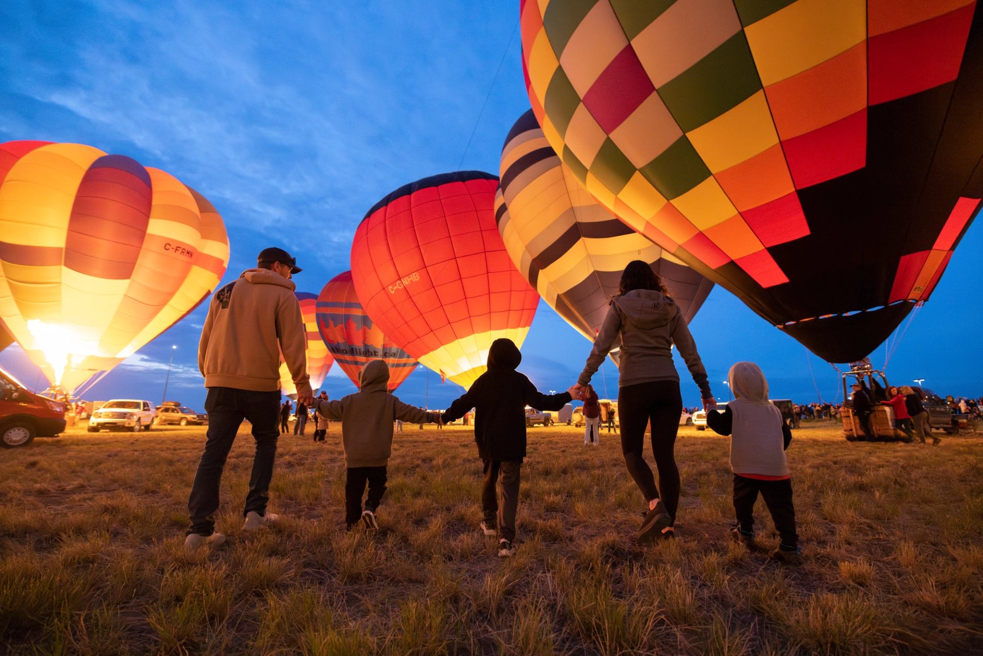 A family walks towards brightly lit hot air balloons at dusk.