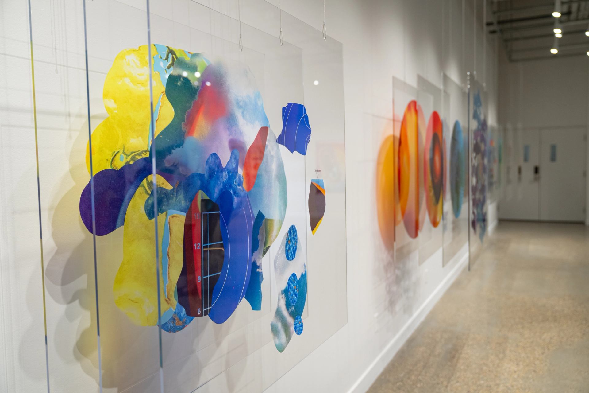 Colourful abstract artworks on transparent panels hang along a bright gallery hallway wall.
