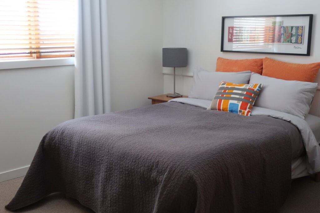 Cozy bedroom with gray quilt, orange pillows, and natural light through window blinds.