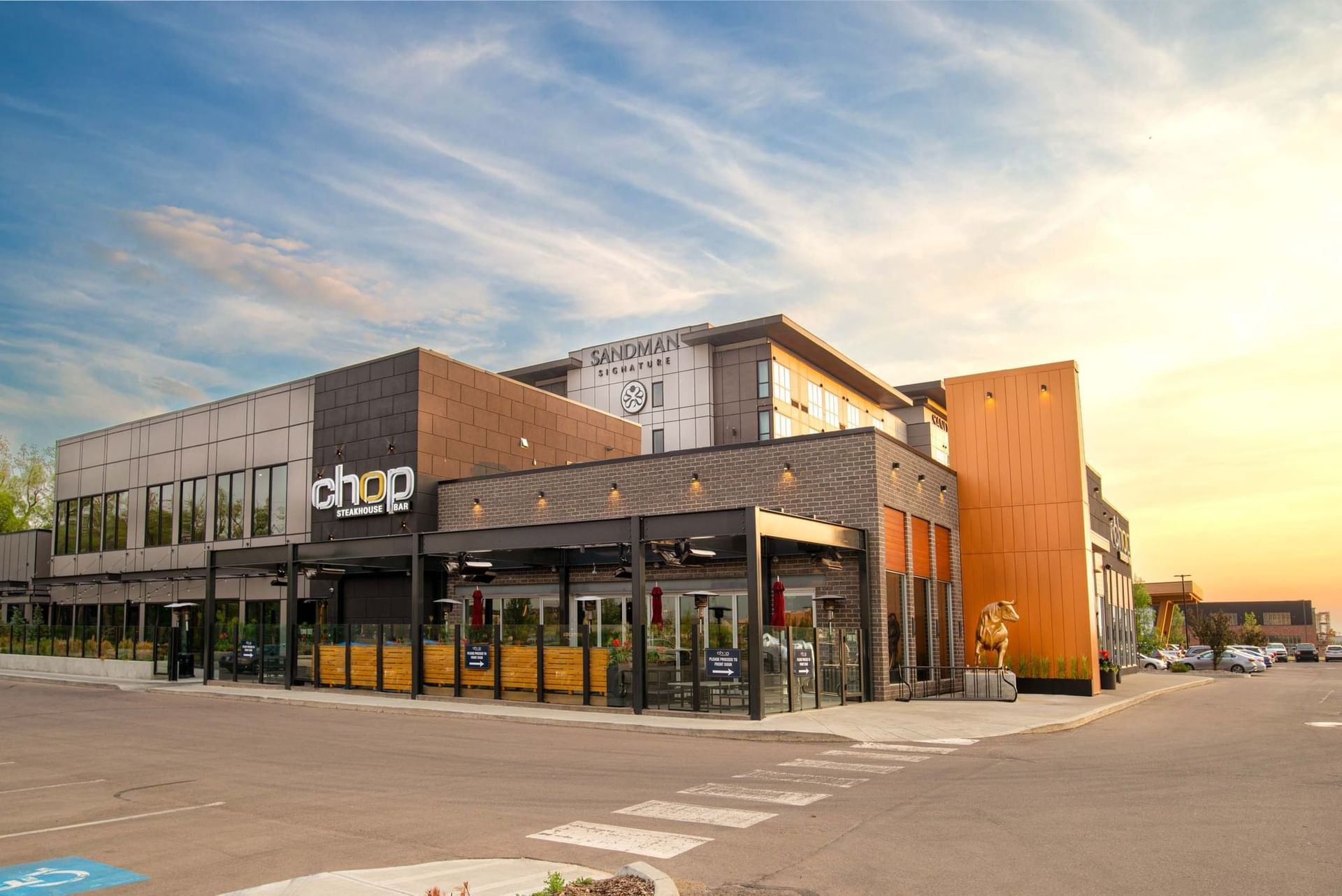 Exterior of Sandman Signature Sherwood Park Hotel featuring modern architecture and Chop Steakhouse & Bar restaurant with outdoor patio seating at sunset.