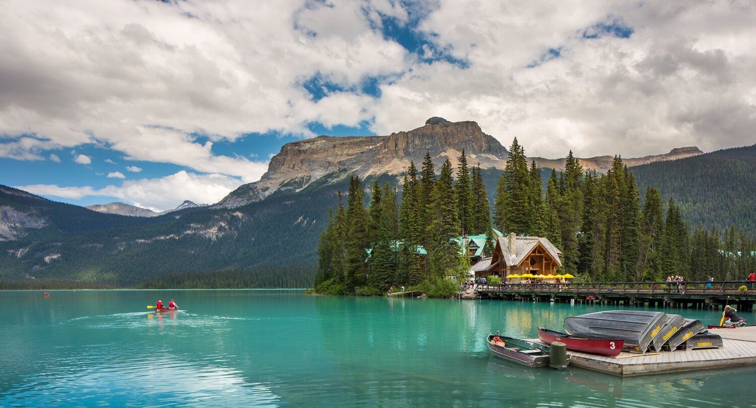 Lake Emerald with boats, lakeside dock, mountains, and a wooden boathouse under clouds.