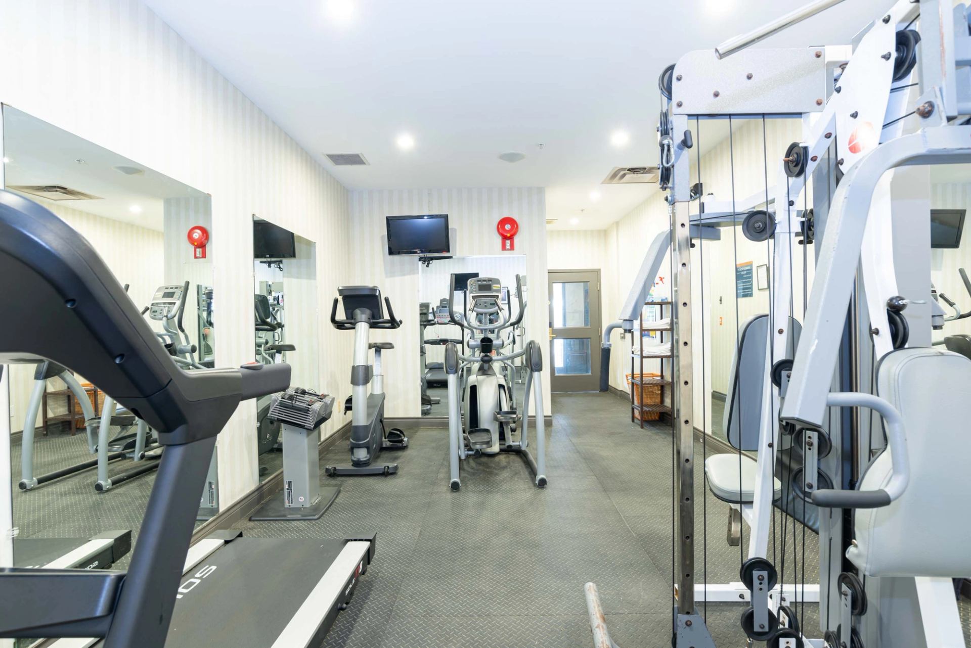 Small fitness room with cardio machines, weight equipment, wall mirrors and mounted television.
