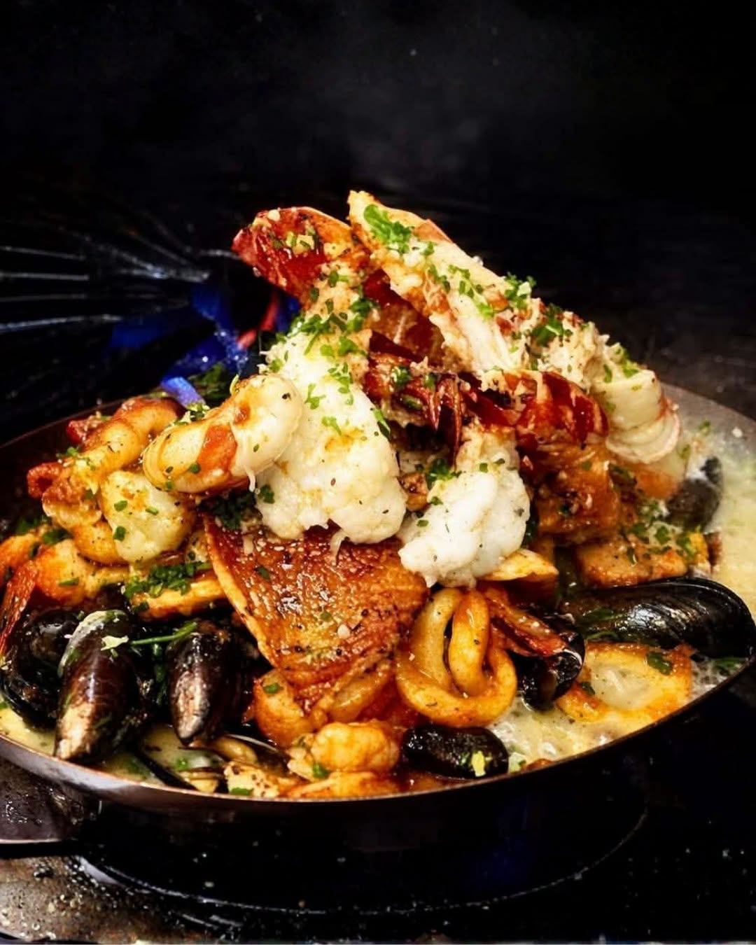 A seafood-rich entrée features lobster, shrimp, mussels, calamari, and fish in a steaming skillet with sauce.