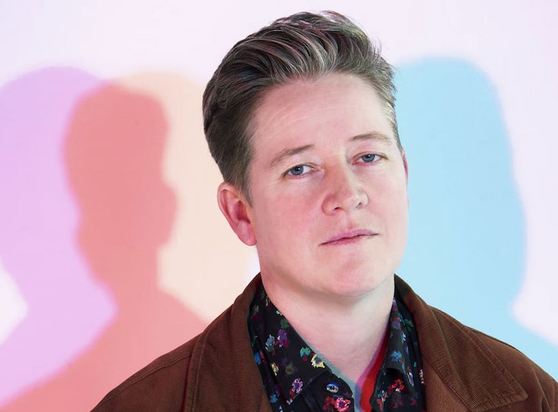 Person with short hair stands before pastel backdrop with overlapping coloured shadows.