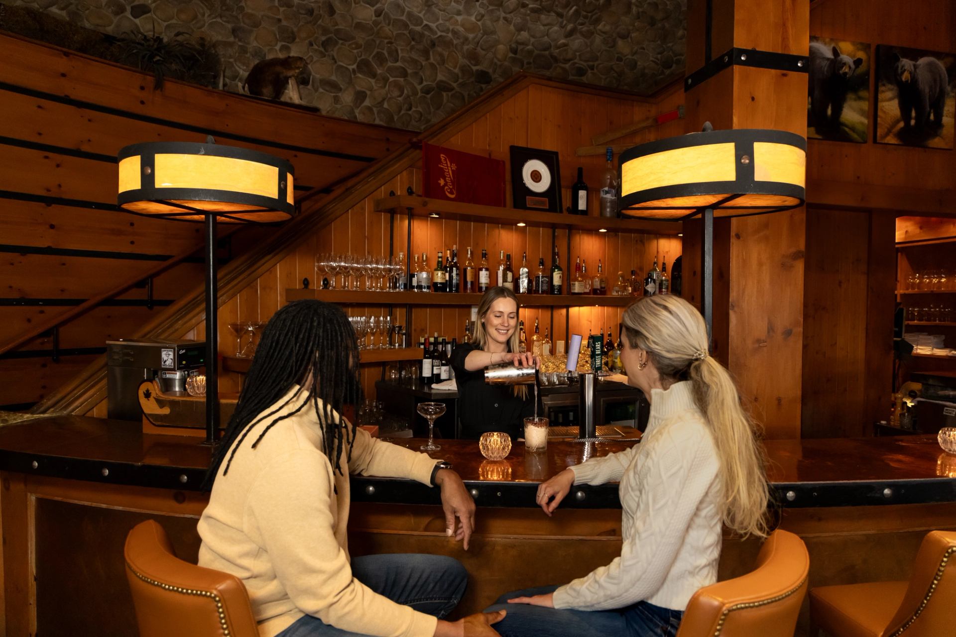 A man and woman sit at the bar while the bartender pours a cocktail. Warm wood interior.