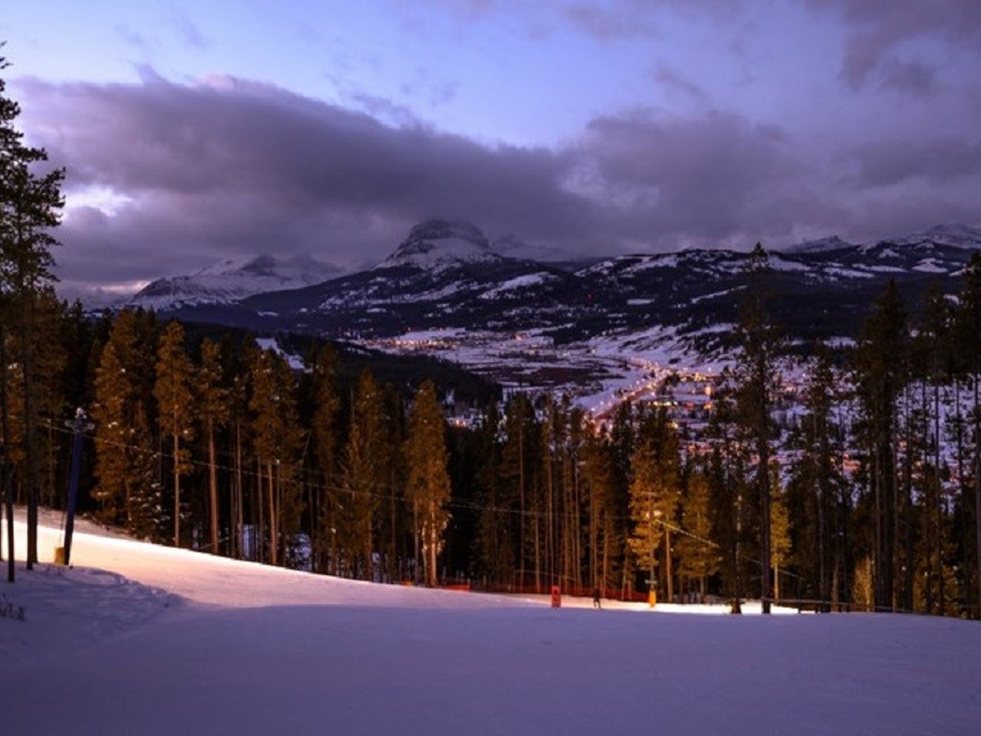 Snow-covered ski slope at Pass Powderkeg with pine trees and mountain backdrop at dusk.
