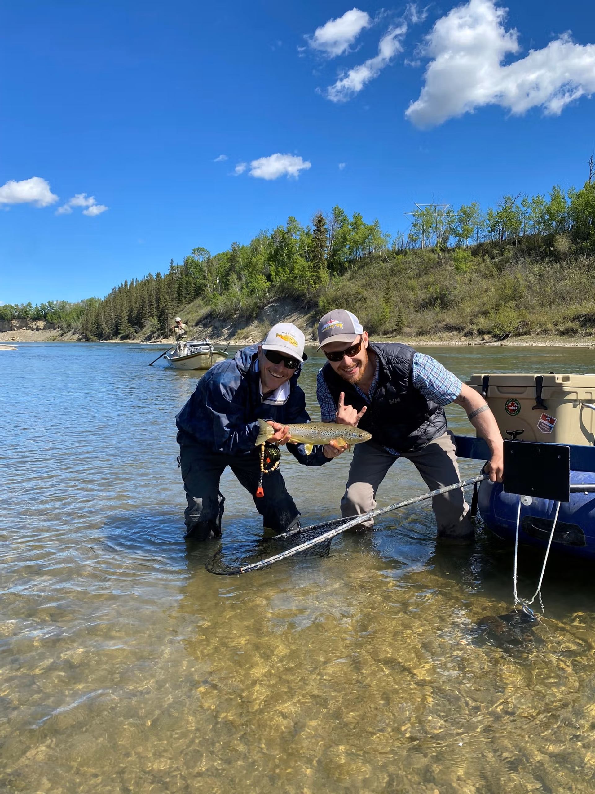 Two people in shallow river water holding a fish beside a blue inflatable boat