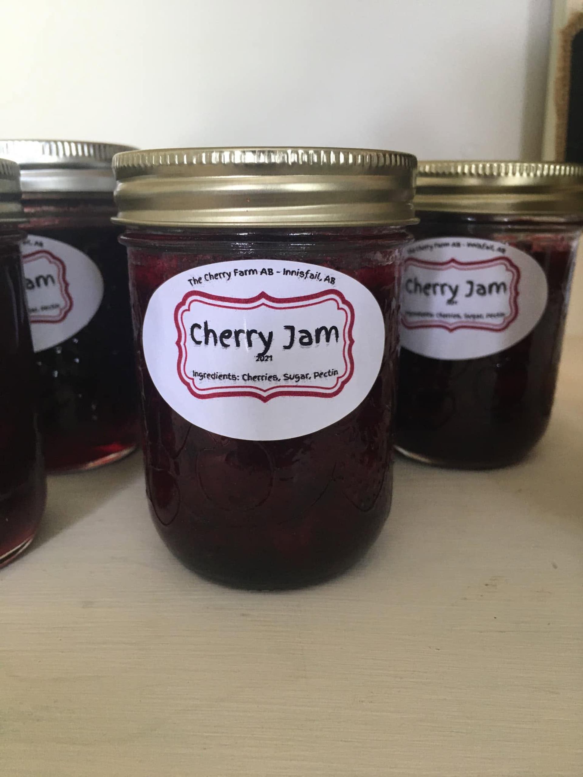 Glass jars filled with dark cherry jam are displayed on a shelf, each sealed with a metal lid.