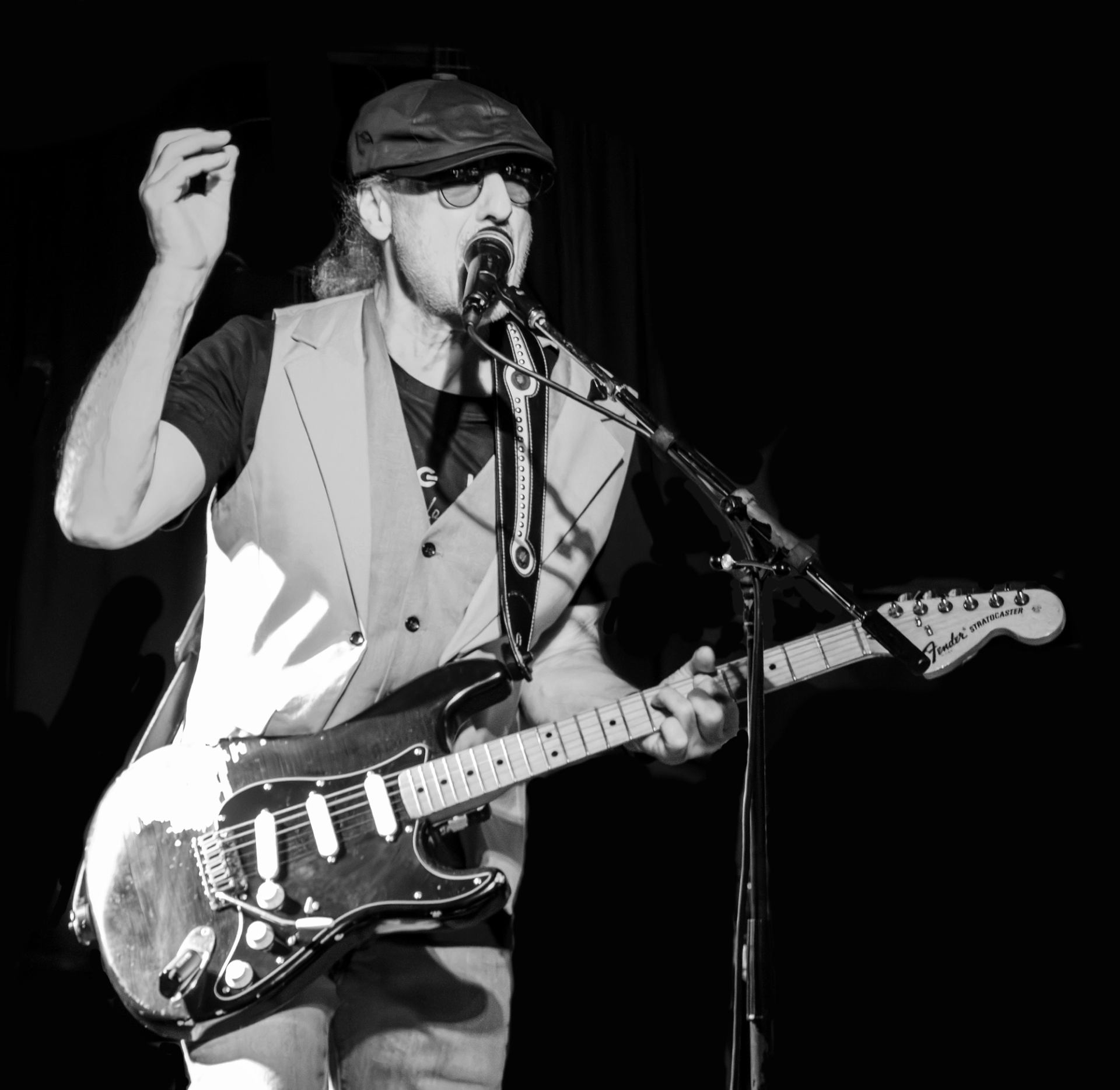 Musician wearing a cap and sunglasses sings while playing an electric guitar.