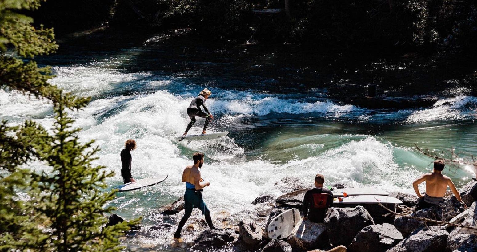 River surfer riding a wave with others watching from the rocky shore.