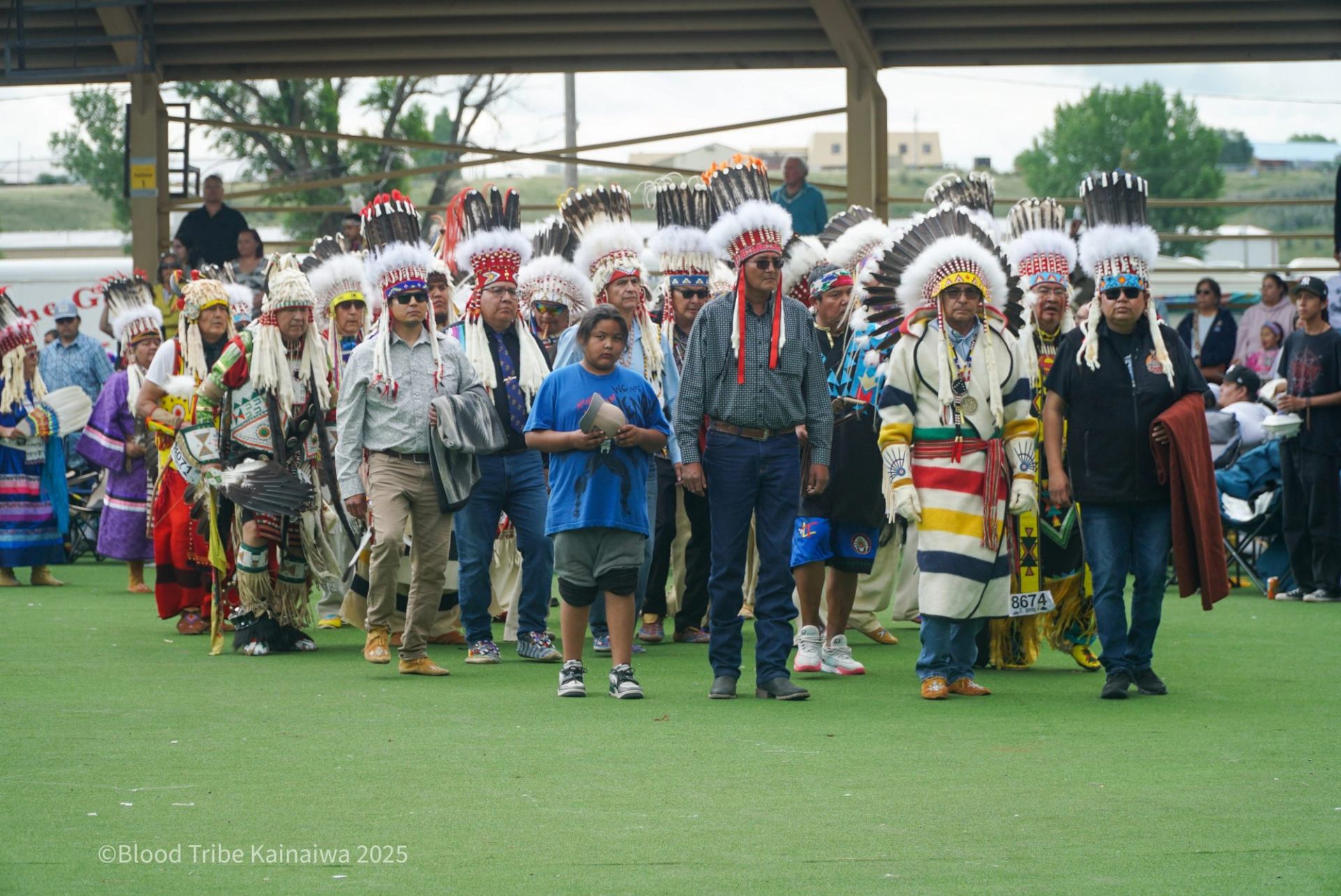 Group of people in regalia and feathered headdresses walking across a covered field.