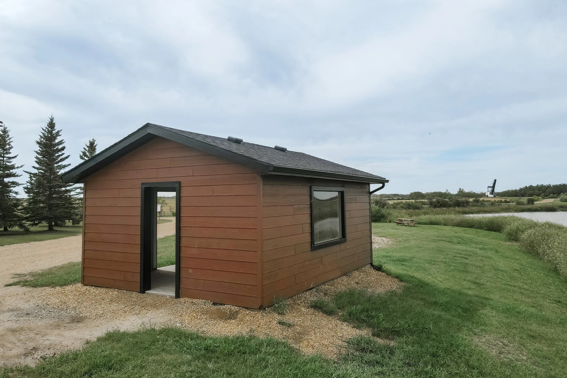 Small brown cabin with open doorway beside gravel path, trees and pond in background.