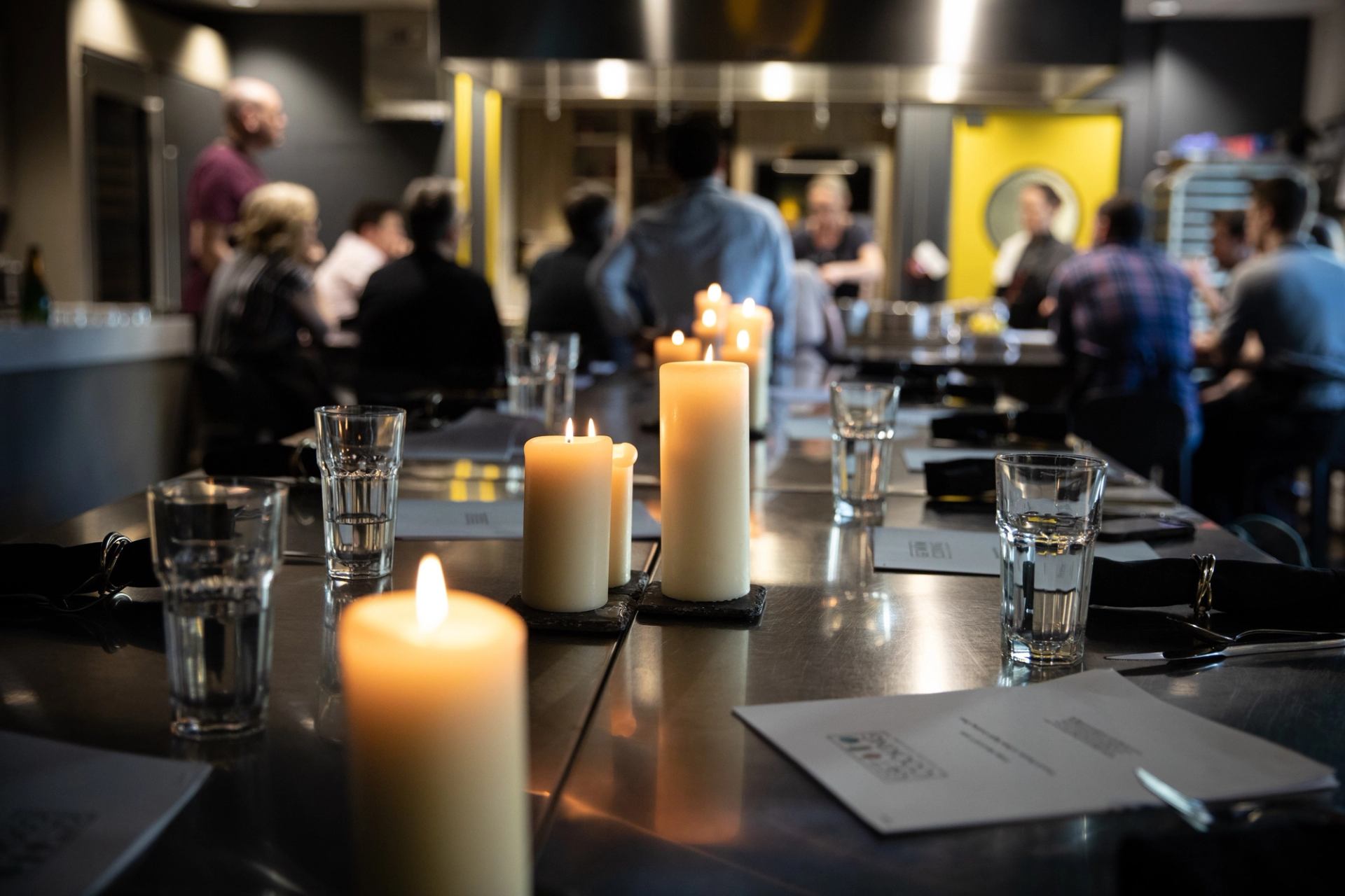 Table setting with candles and drinks, with people in a kitchen workshop background.