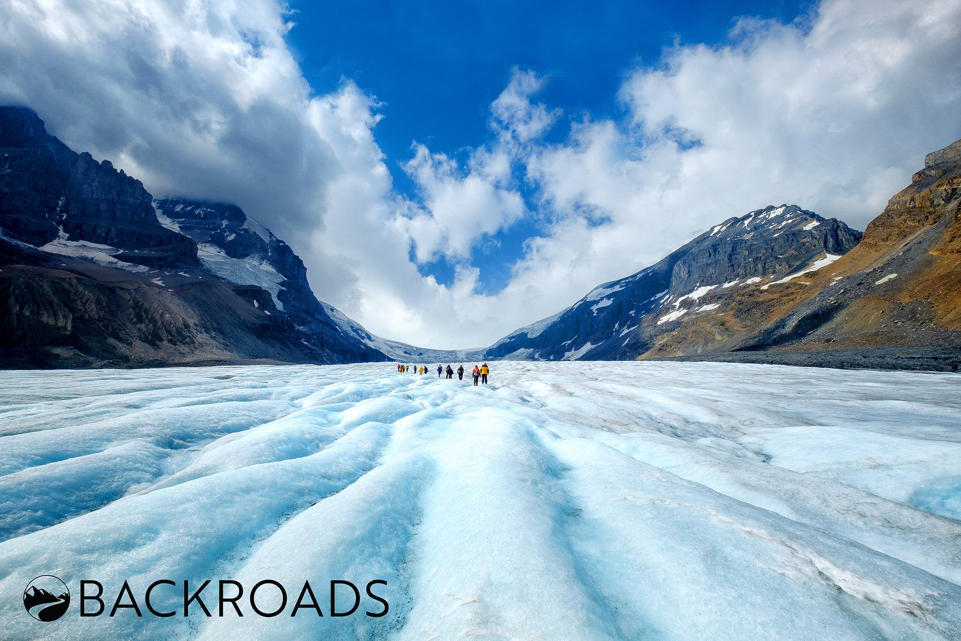 Group walking on glacier with blue ice, snowy peaks, and cloudy sky in the background.