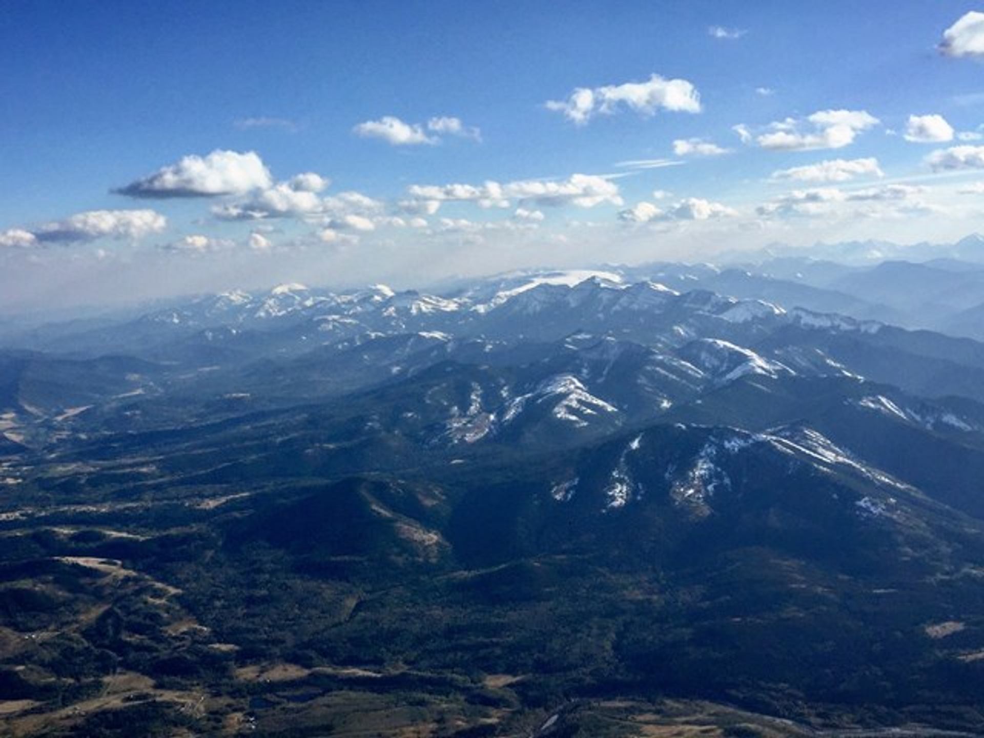 Snow-capped mountain range under a bright blue sky with scattered clouds, viewed from above.