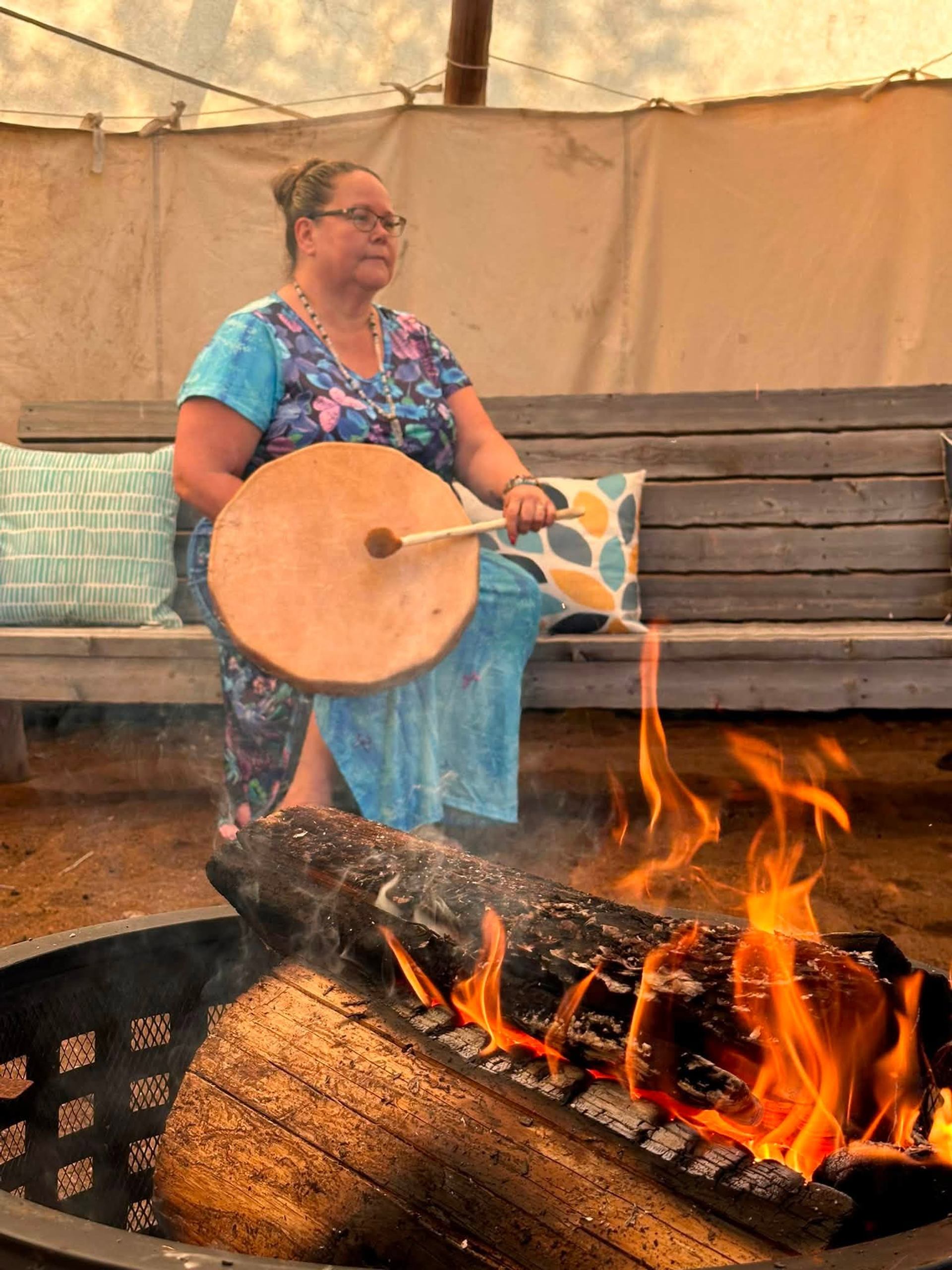 Person in blue dress holding hand drum near open fire inside tented space.