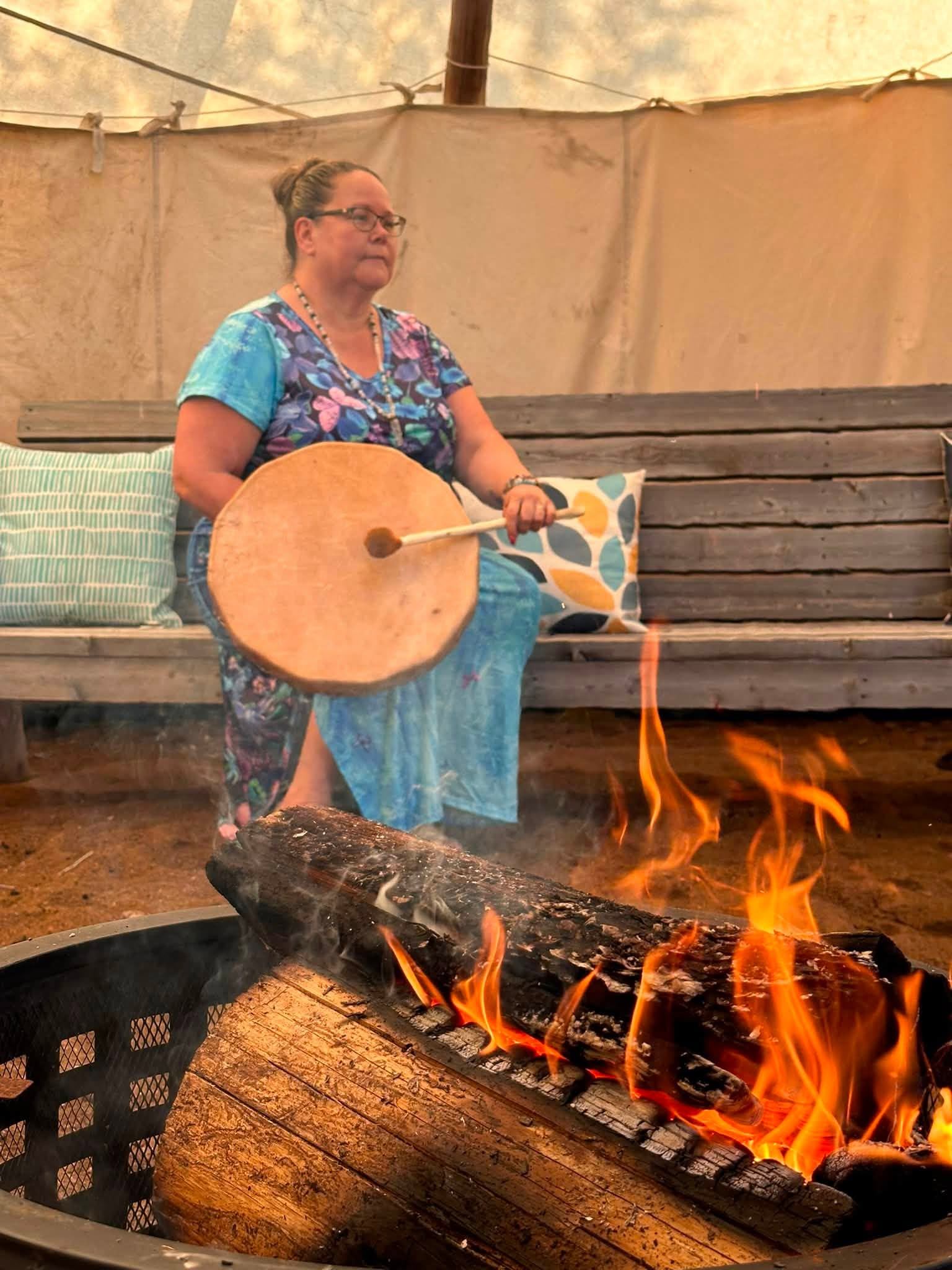Person in blue dress holding hand drum near open fire inside tented space.