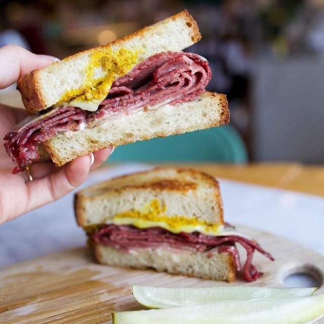 Hand holds sliced meat sandwich; other half and pickle spear on cutting board.