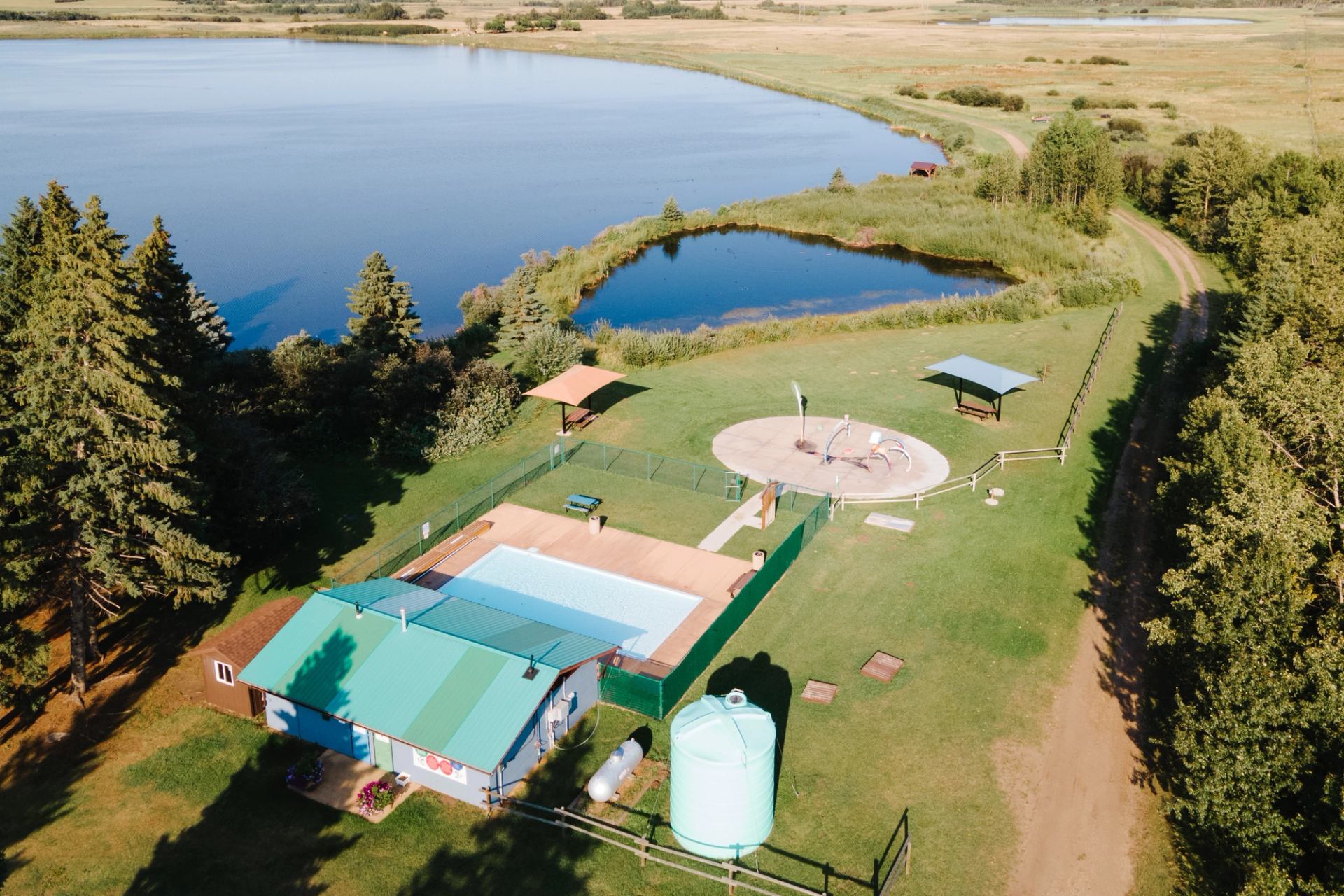 Aerial view of lakeside park with outdoor pool, splash pad and surrounding trees.