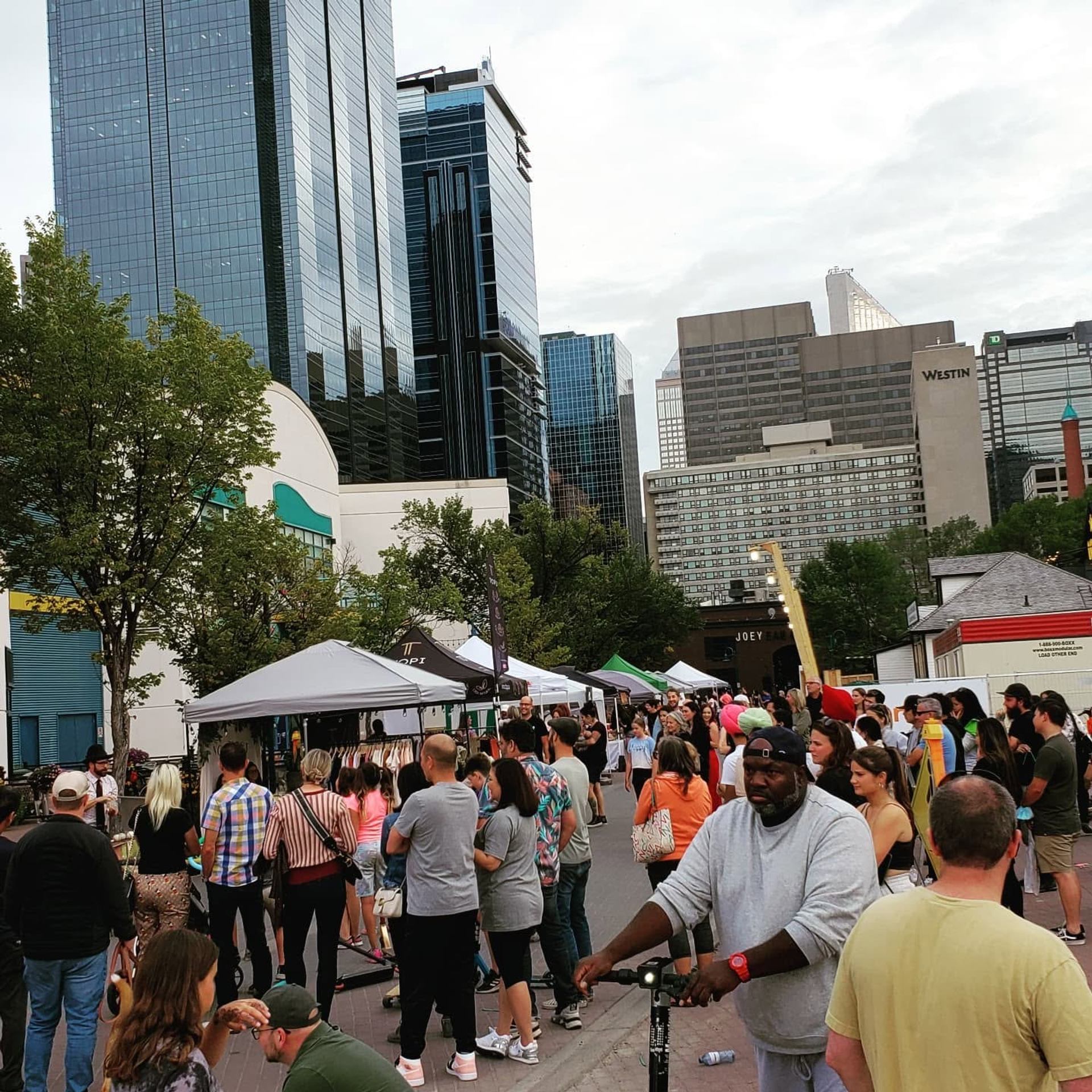 Crowd browsing outdoor market stalls on city street with tall office buildings behind.