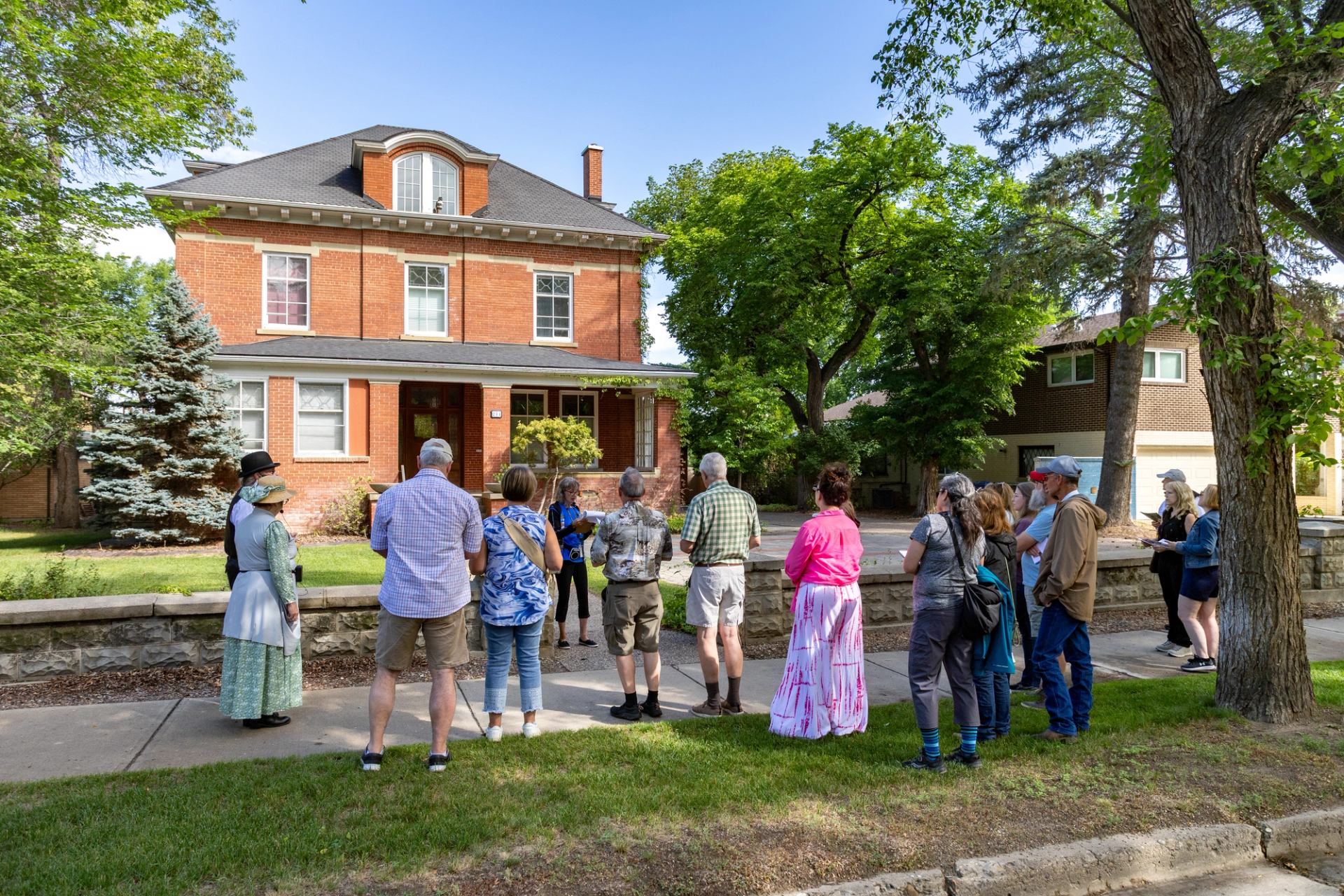 Group of people on sidewalk listening to guide outside large red brick house.