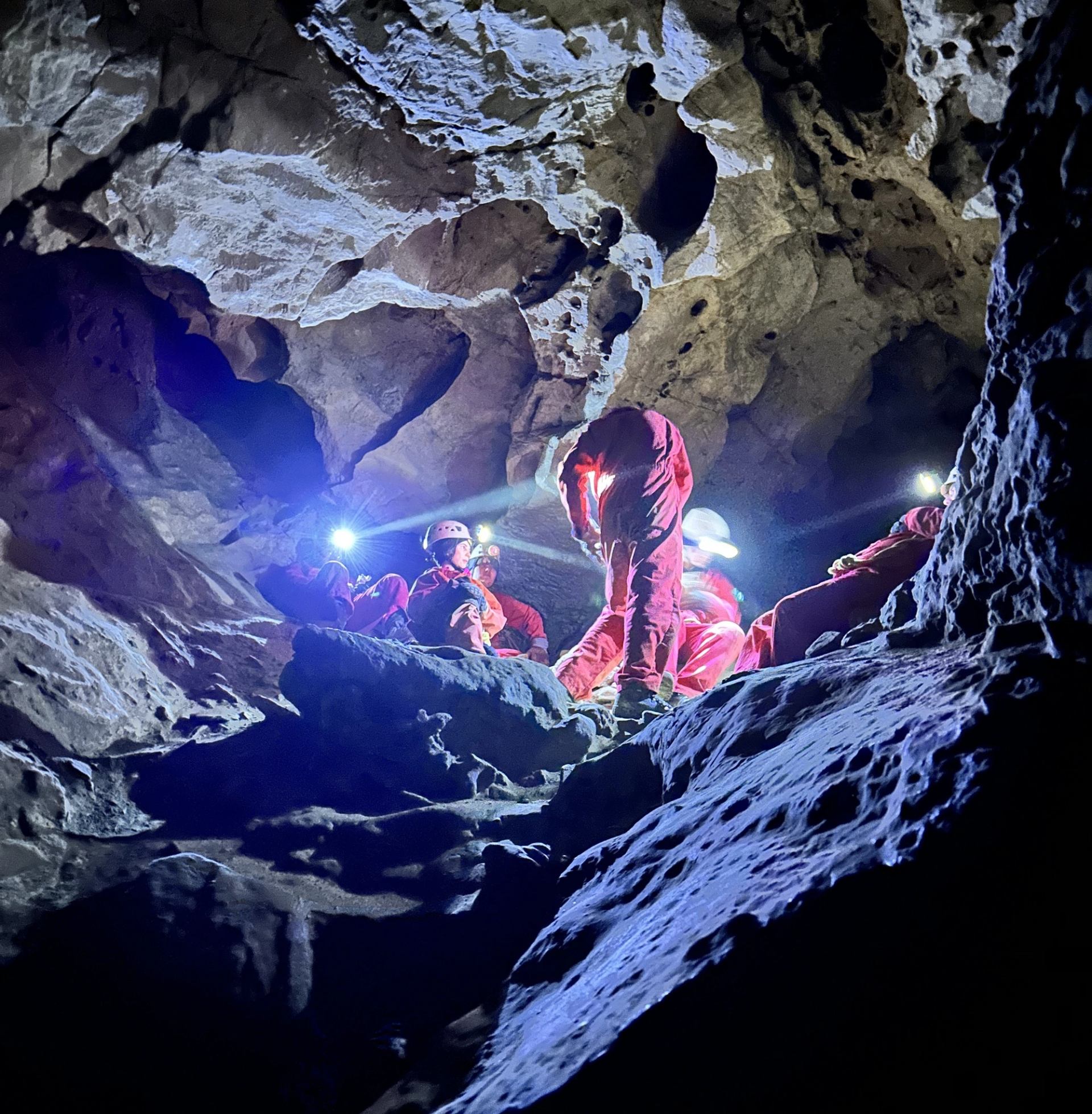 Cavers wearing red suits and headlamps gather among rocks inside a dark cave.
