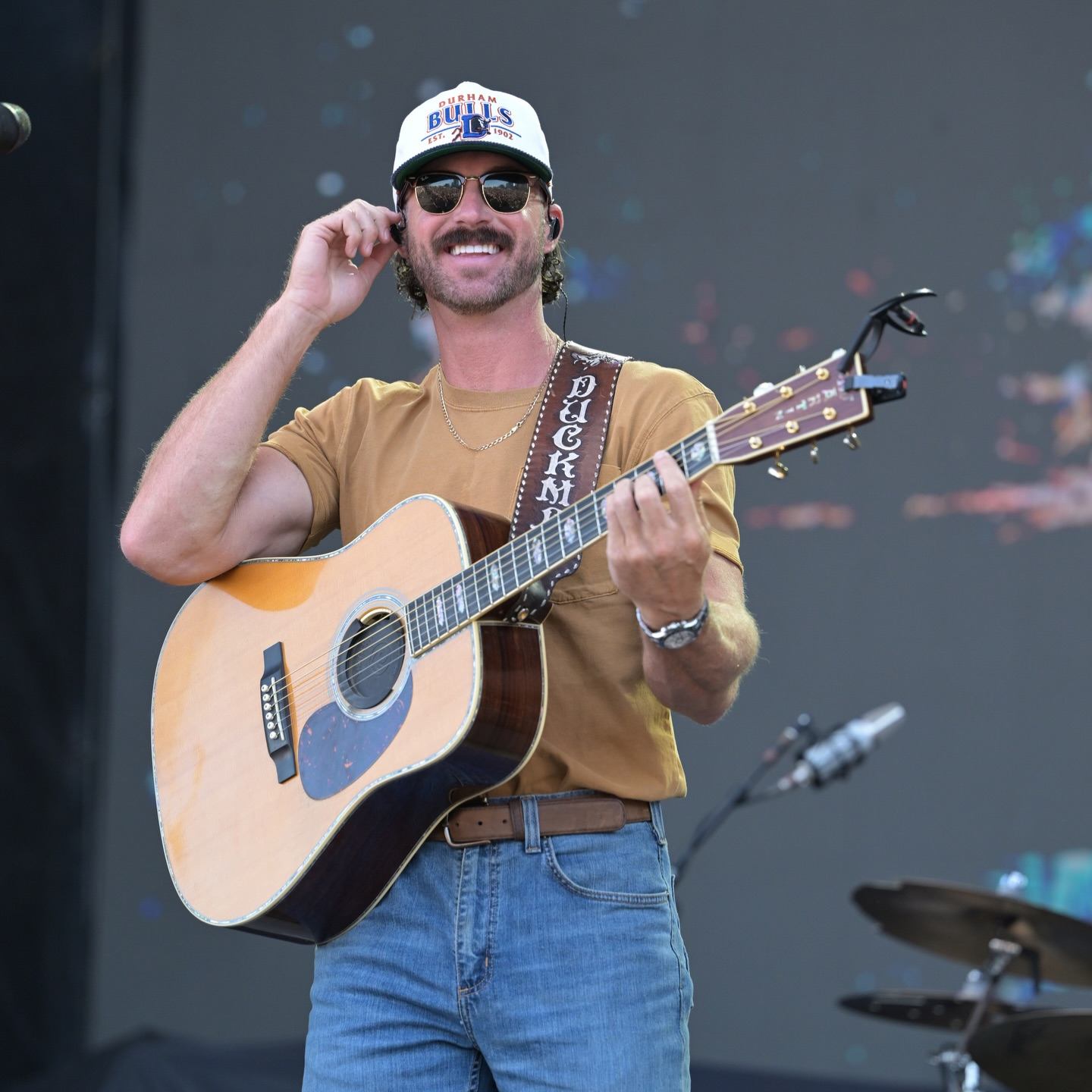 Guitarist wearing sunglasses and a Durham Bulls cap performs onstage with microphone and drums.