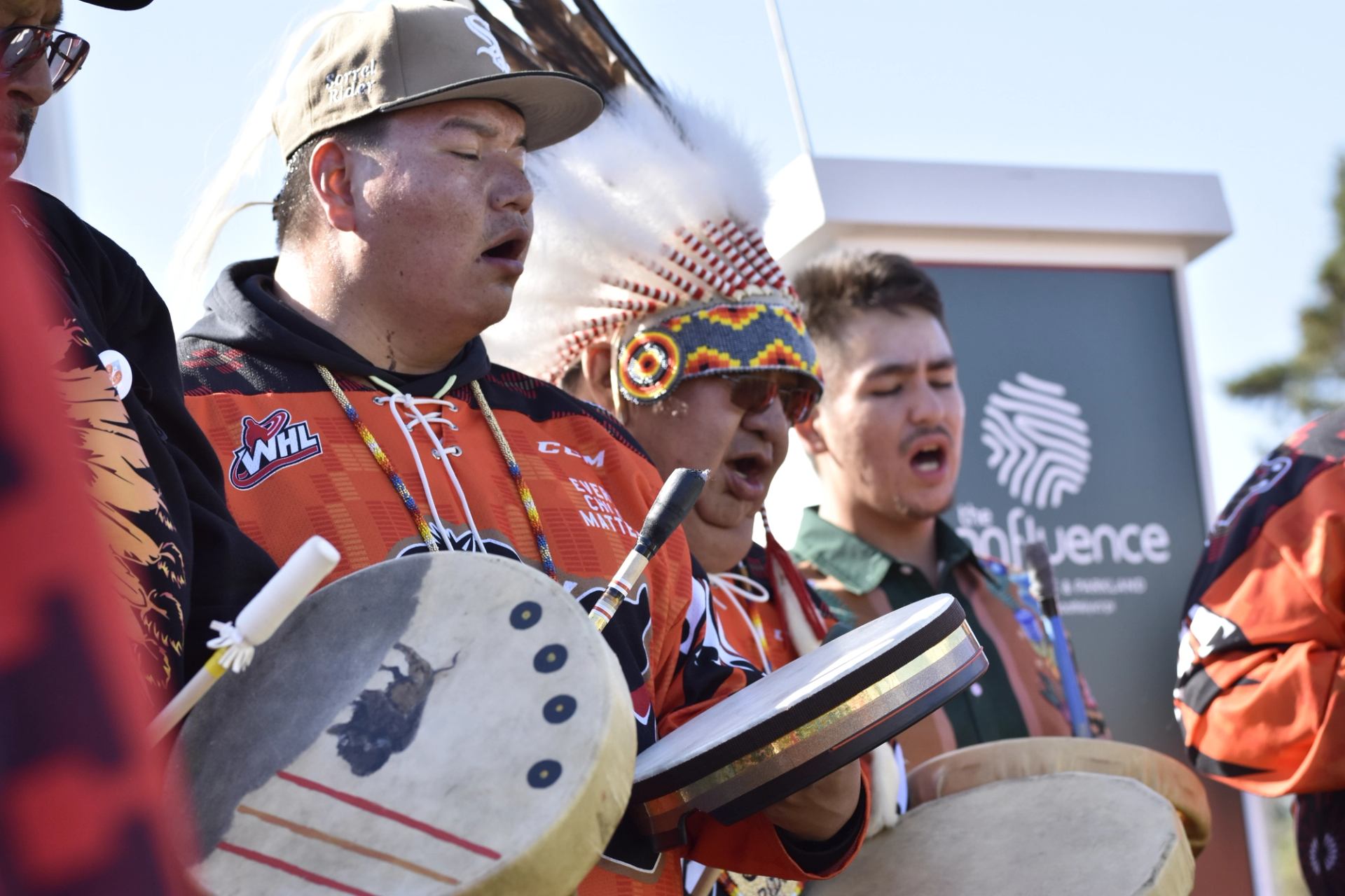 Indigenous drummers in colourful regalia sing together while playing hand drums outdoors.