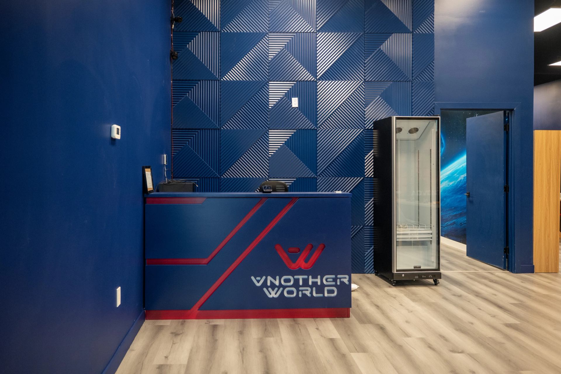 Vnother World reception desk beside a glass-front refrigerator and open blue door.