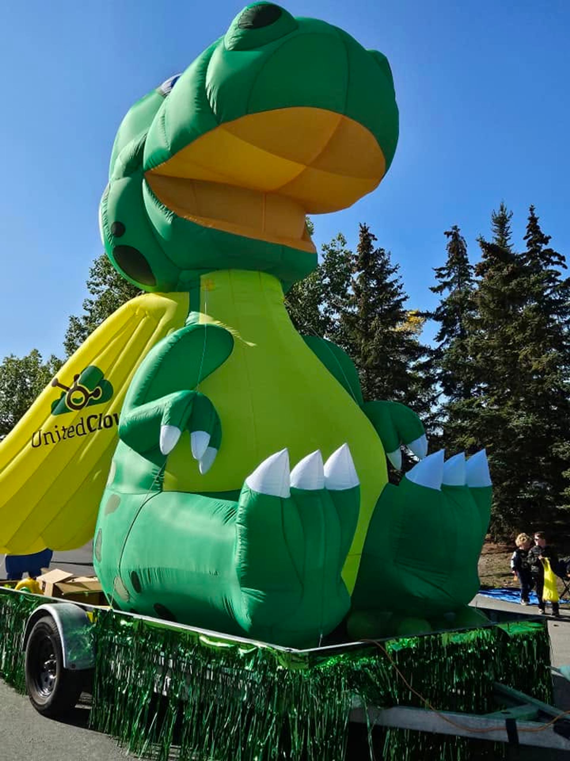 Large inflatable green dinosaur on parade float with United Clover banner and trees behind.