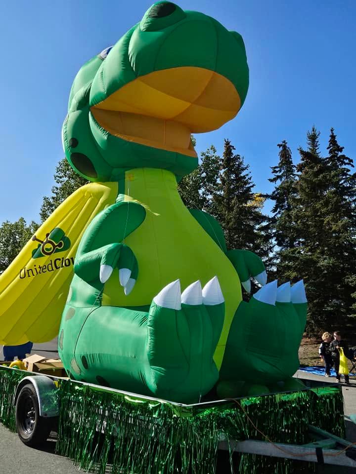 Large inflatable green dinosaur on parade float with United Clover banner and trees behind.