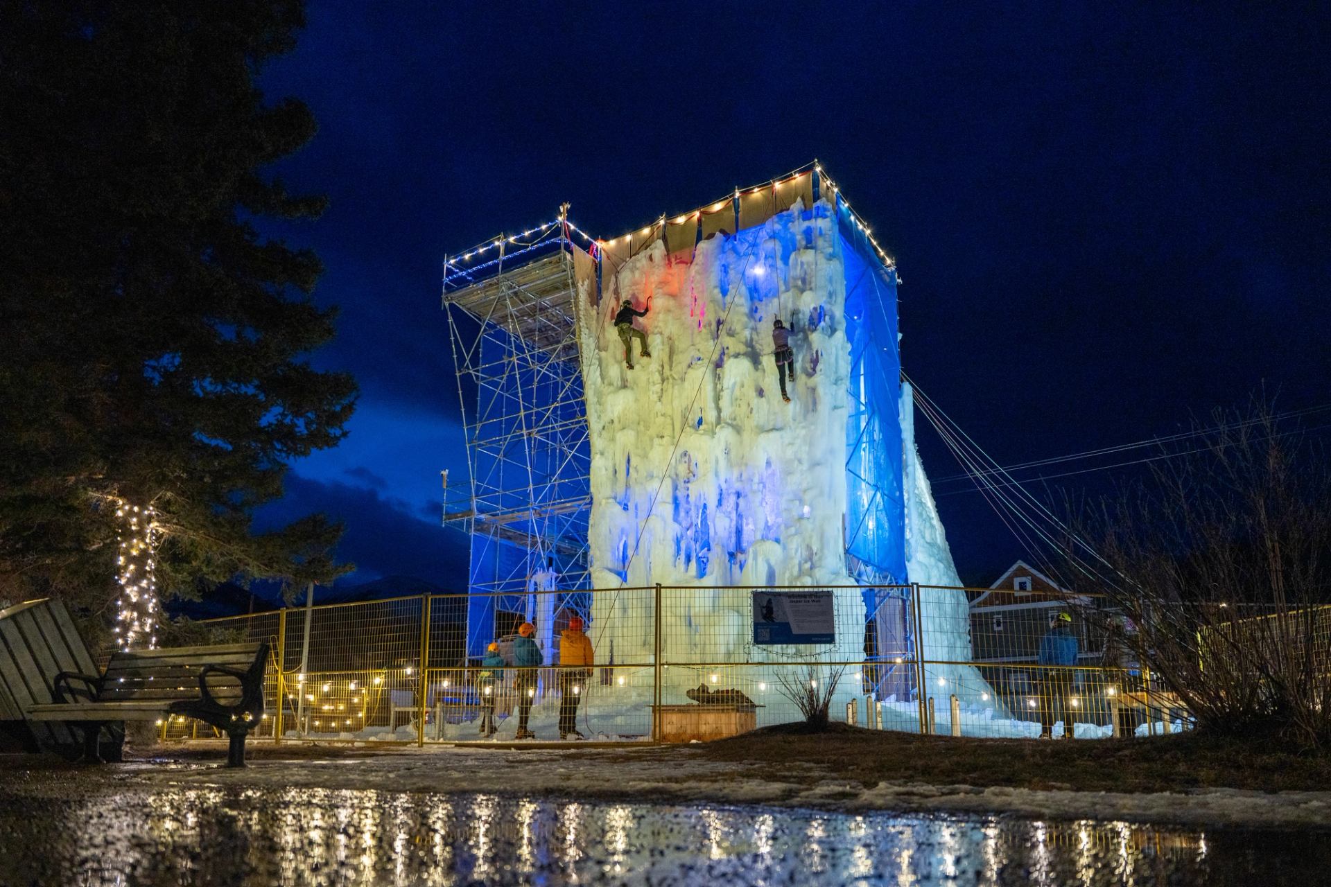 Climbers scale lit-up ice wall at night with ropes under a dark sky.