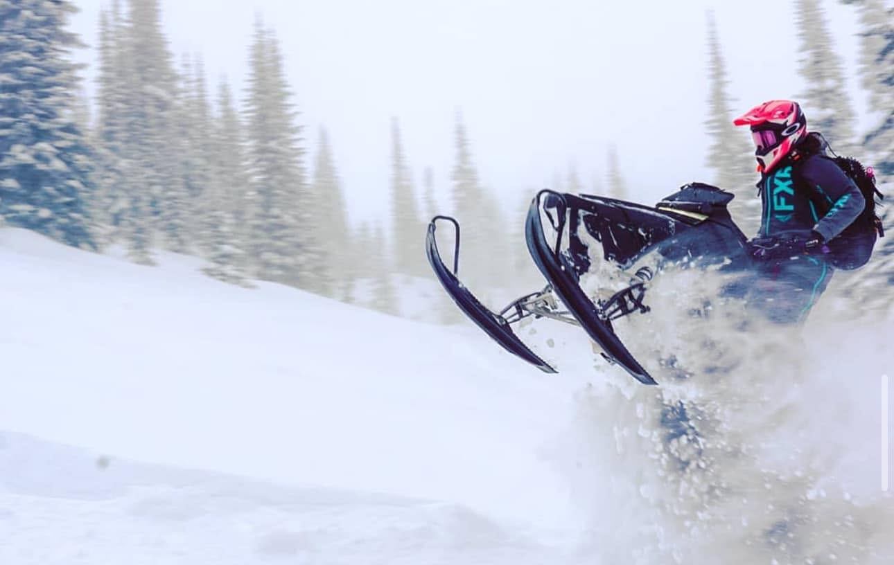 Snowmobiler lifts the front skis while riding through deep snow among evergreens.