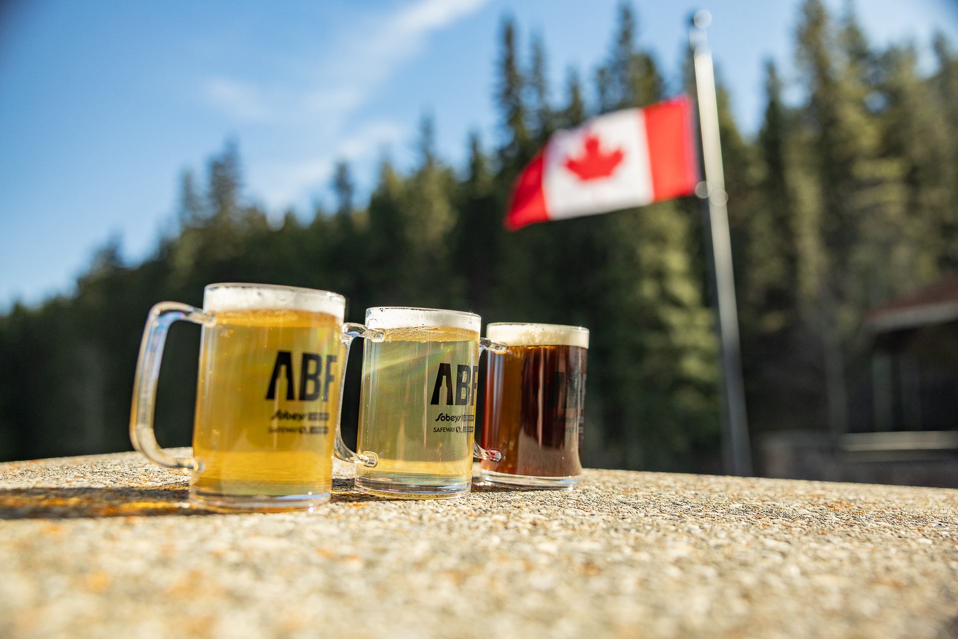 Three ABF beer mugs on a stone surface with blurred Canadian flag behind.
